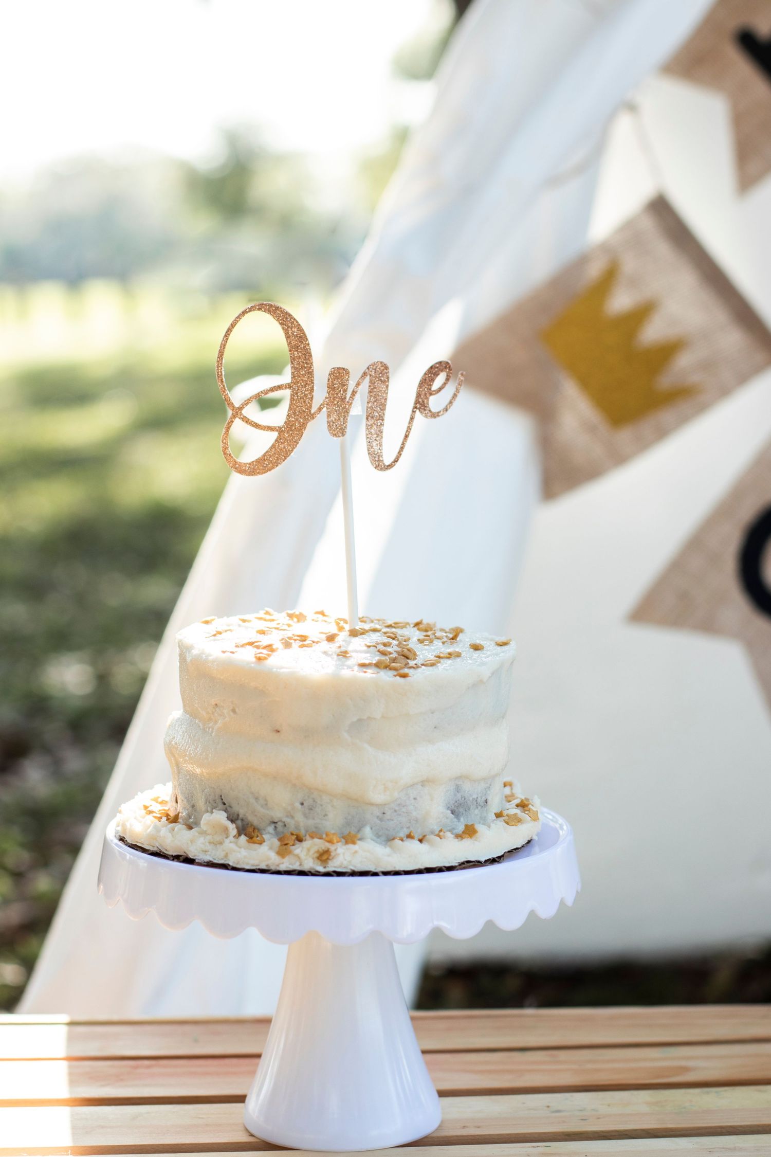 White birthday cake with gold 'One' topper displayed on pedestal against teepee backdrop.