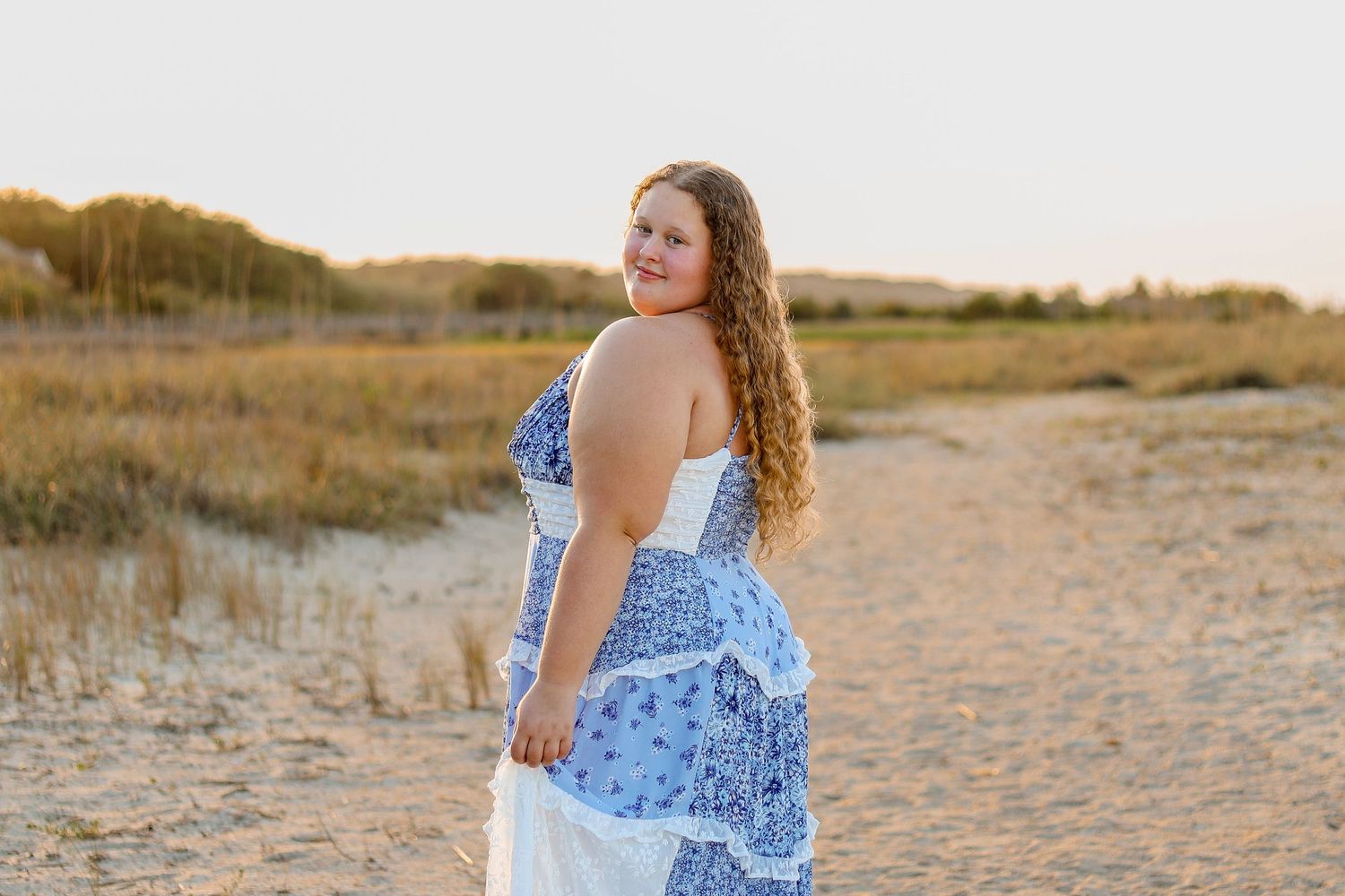 A person in a blue and white patterned summer dress poses at sunset on a sandy beach path.