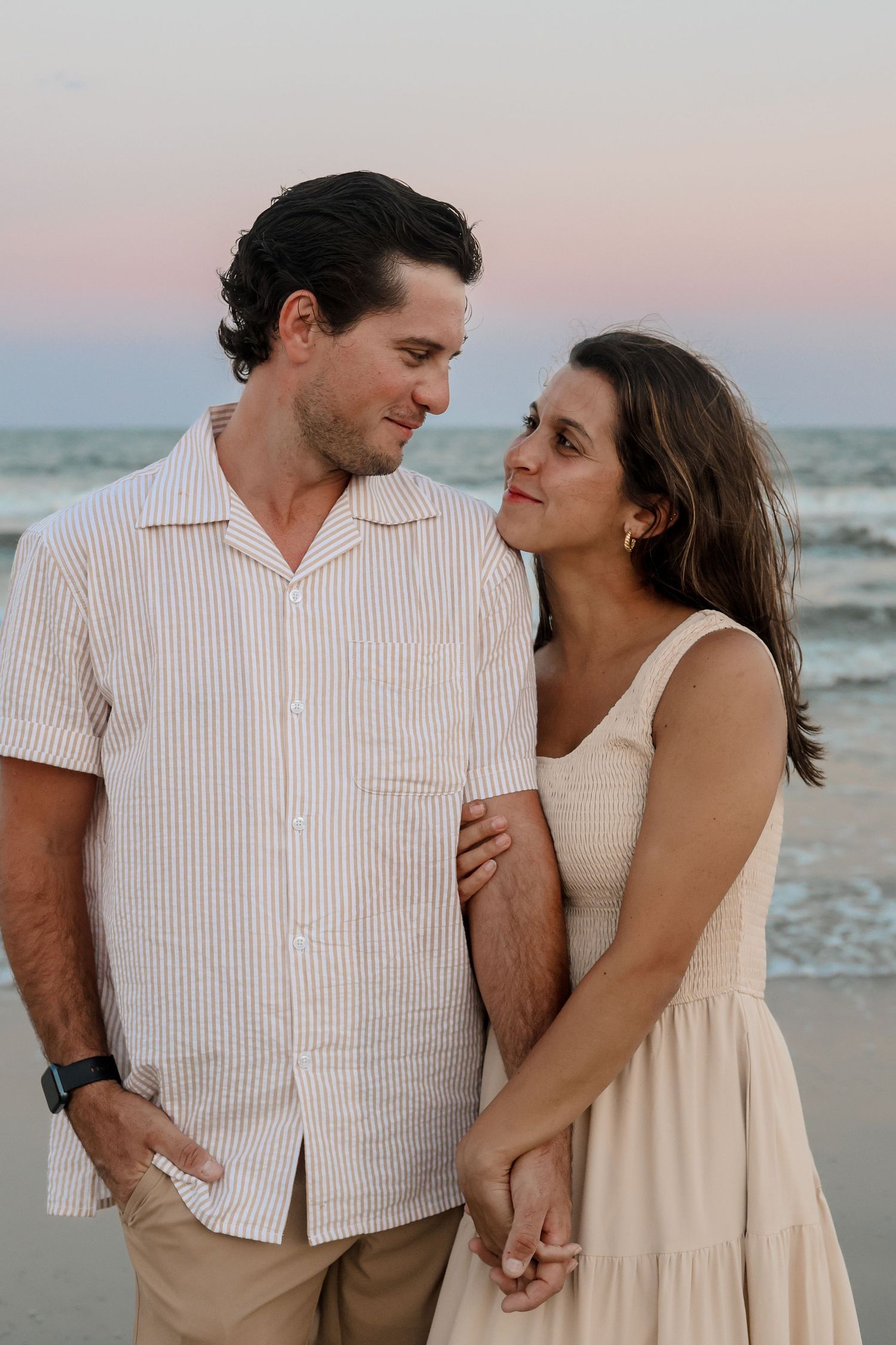 A couple shares romantic moments at sunset on the beach wearing coordinating cream and white outfits.