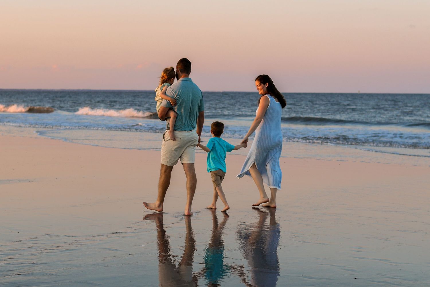Family members holding hands while walking through shallow ocean waves at sunset.