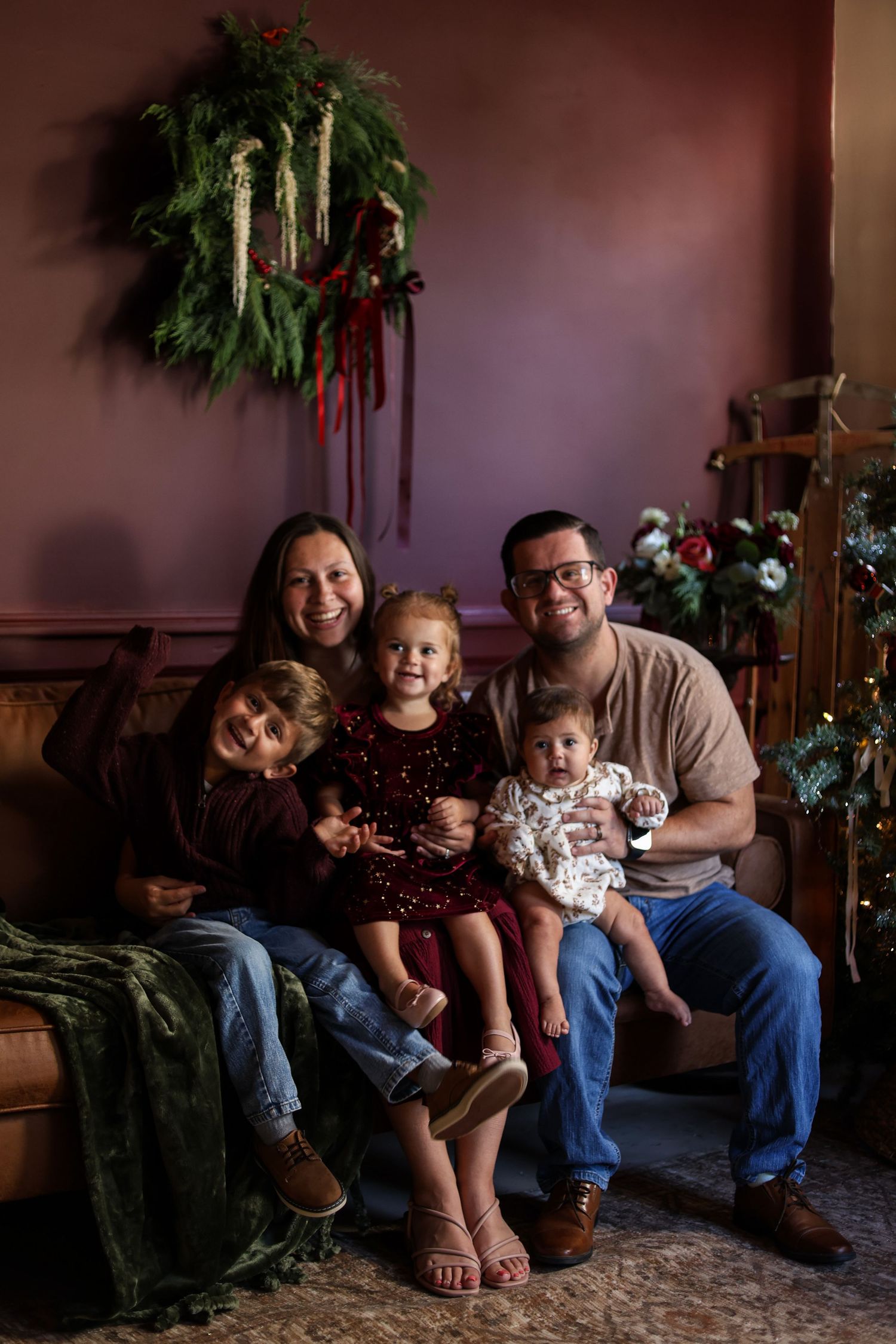 Smiling family of five poses on a couch in a cozy holiday setting with a evergreen wreath and Christmas greenery against a burgundy wall.