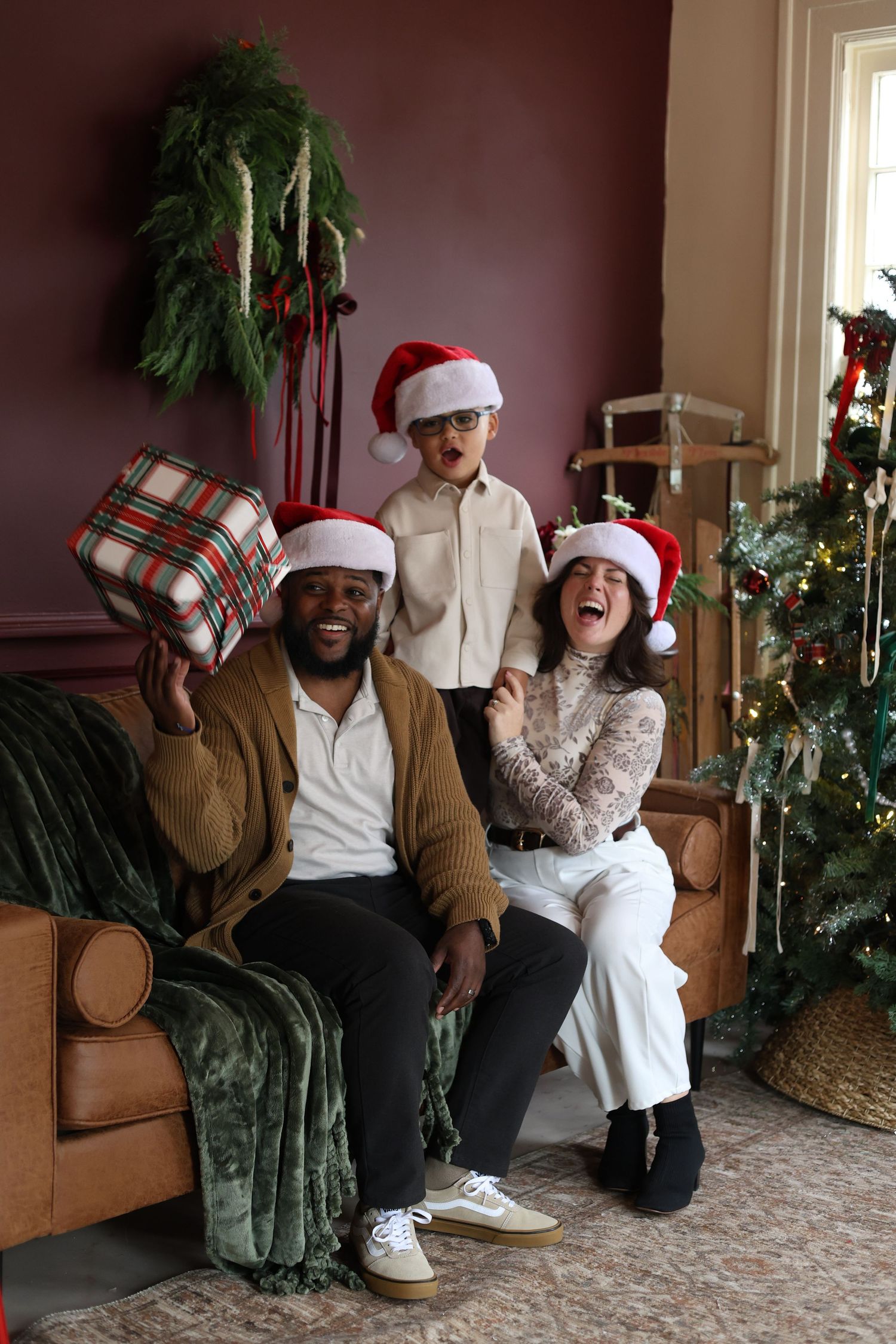 Family in Santa hats laughs on a leather sofa while a man holds up a plaid wrapped gift beside a decorated Christmas tree.