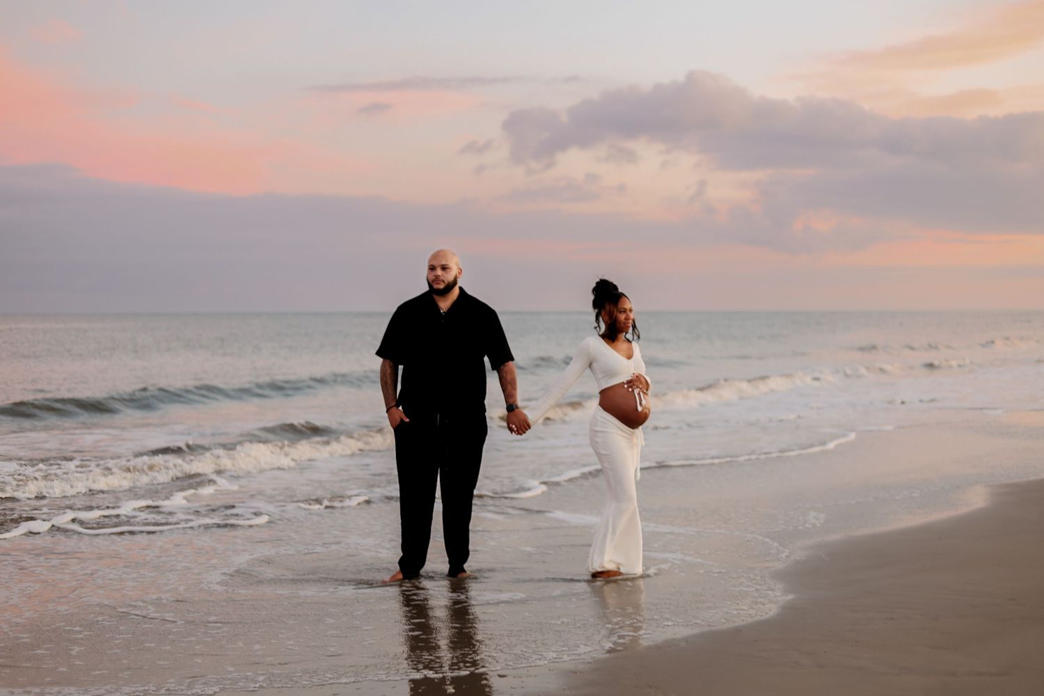 A couple walks along a beach at sunset with waves crashing against the shore in the background.