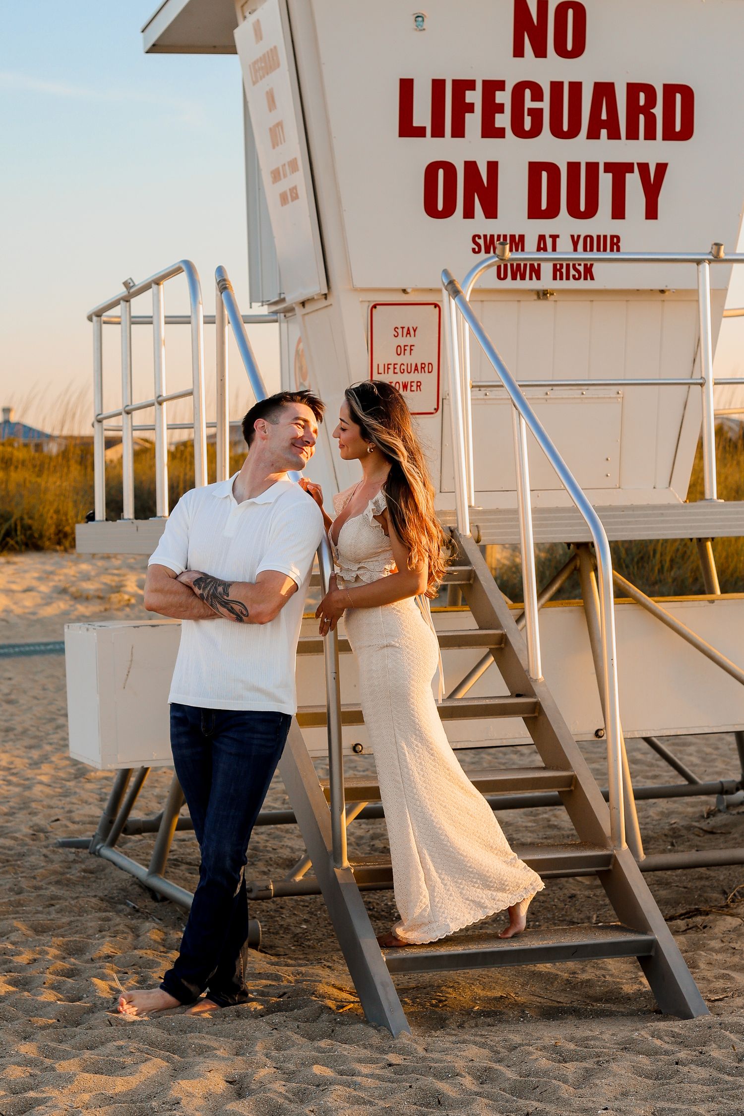 Couple in white outfits standing on lifeguard tower steps at beach during sunset.