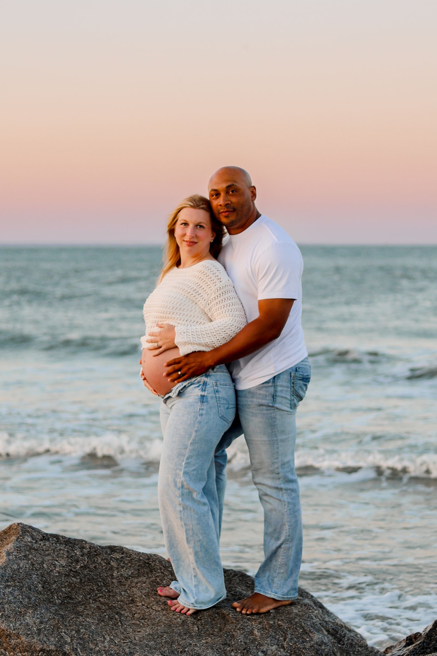 An expectant couple embracing on beach rocks during a pink sunset.