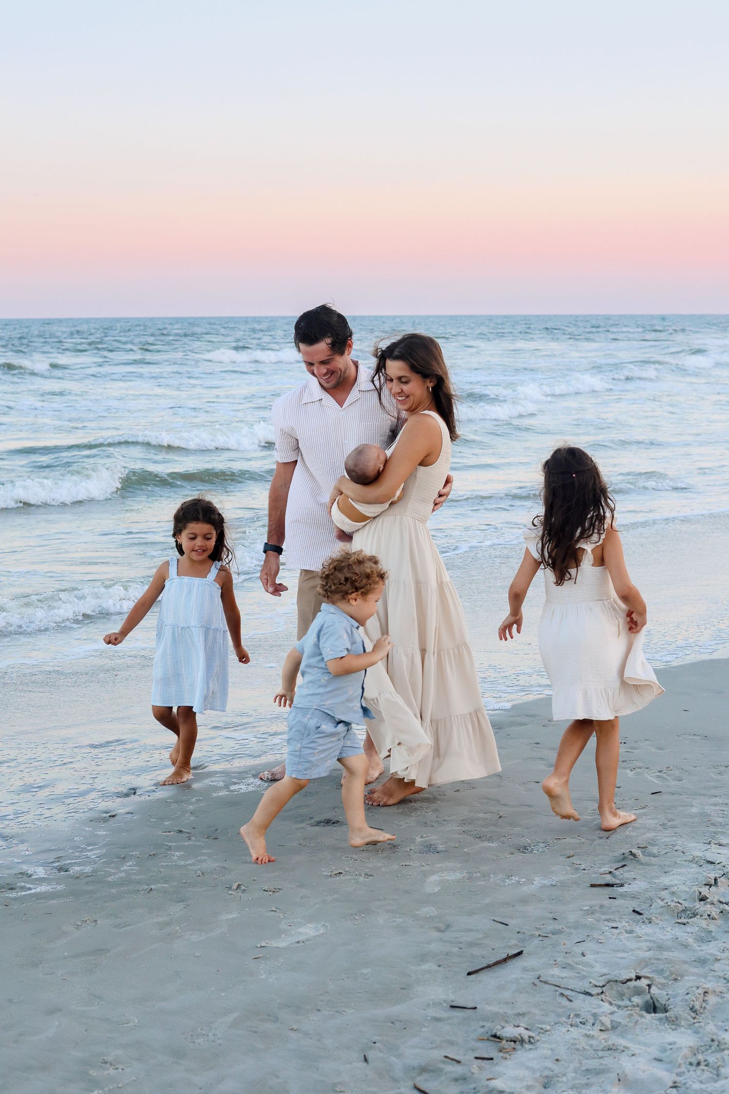 A family walks together barefoot along the beach at sunset with gentle waves rolling in on the shore.