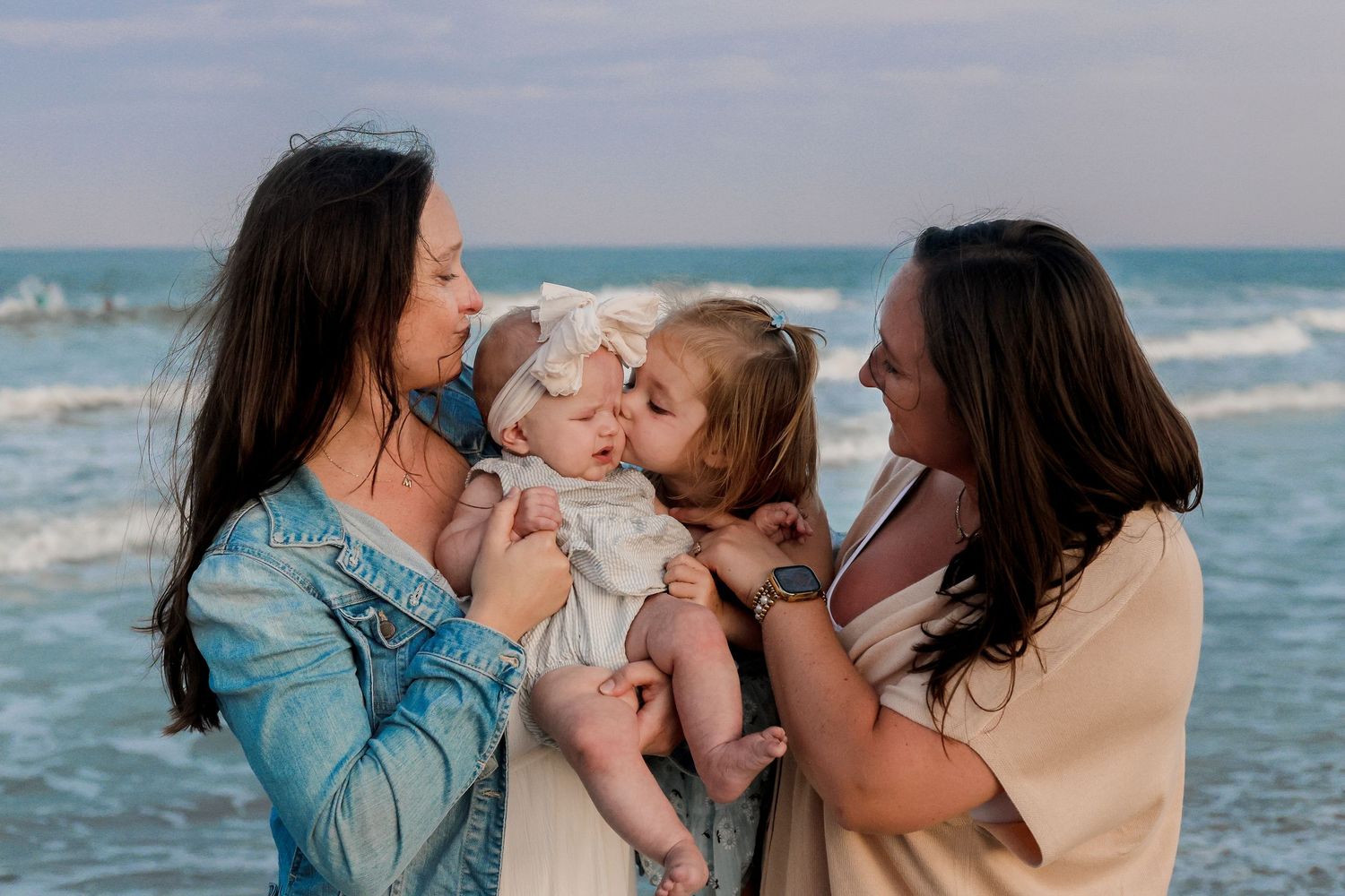 Family enjoying tender moment together at beach with waves and blue ocean in background during sunset.