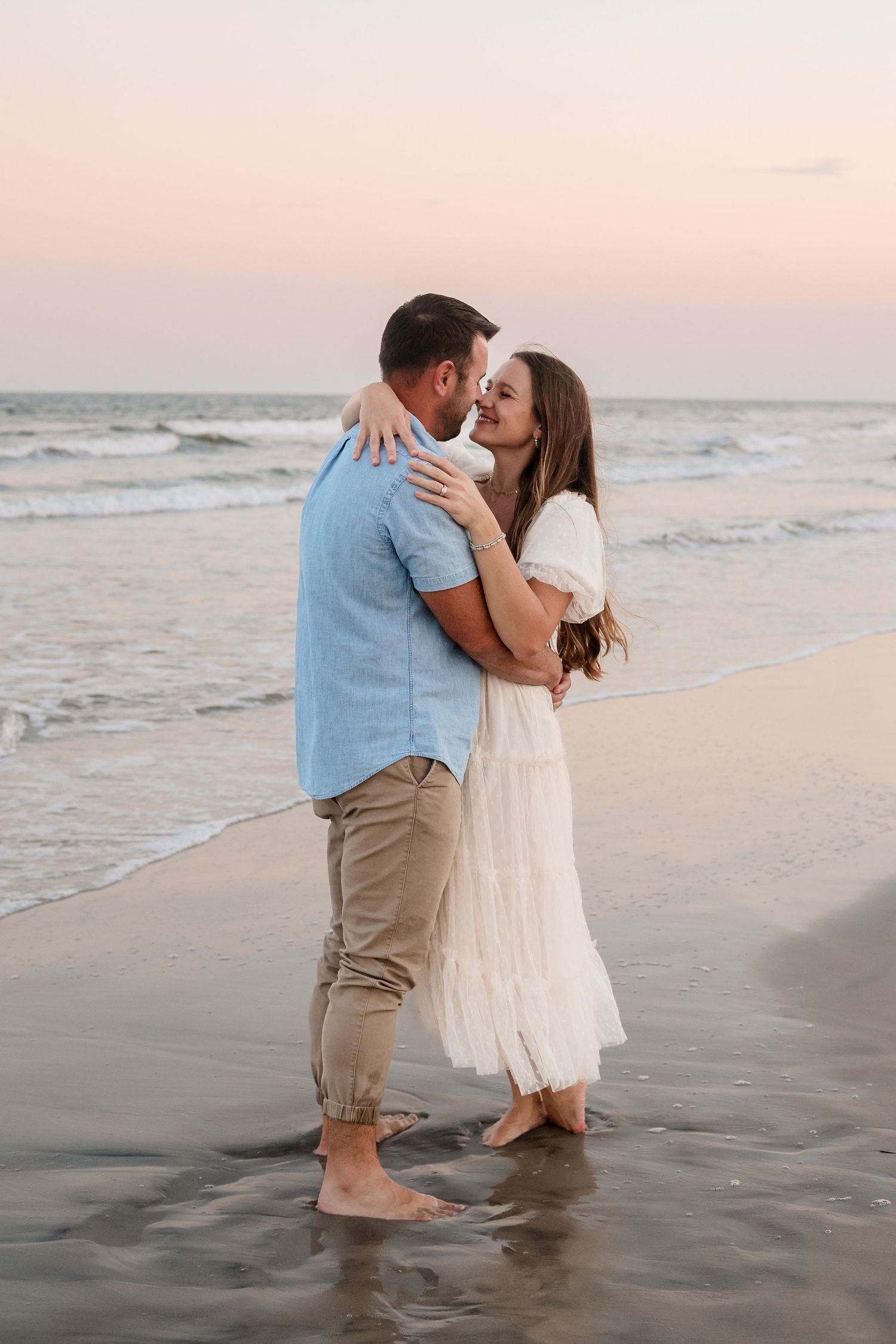 A couple embraces on a beach at sunset with ocean waves in the background.
