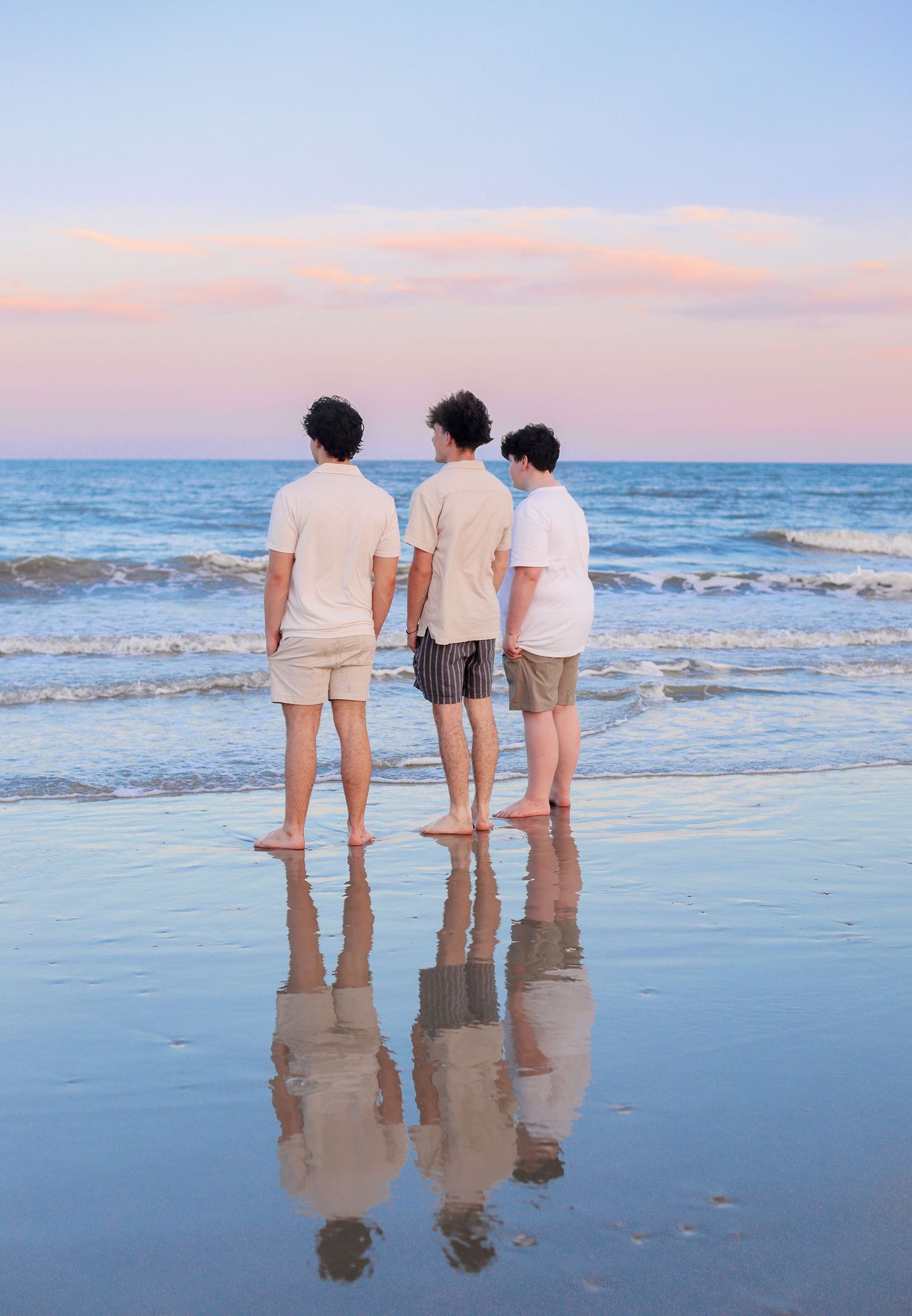 Three people stand together at the edge of the ocean during sunset, their reflections visible in the wet sand.
