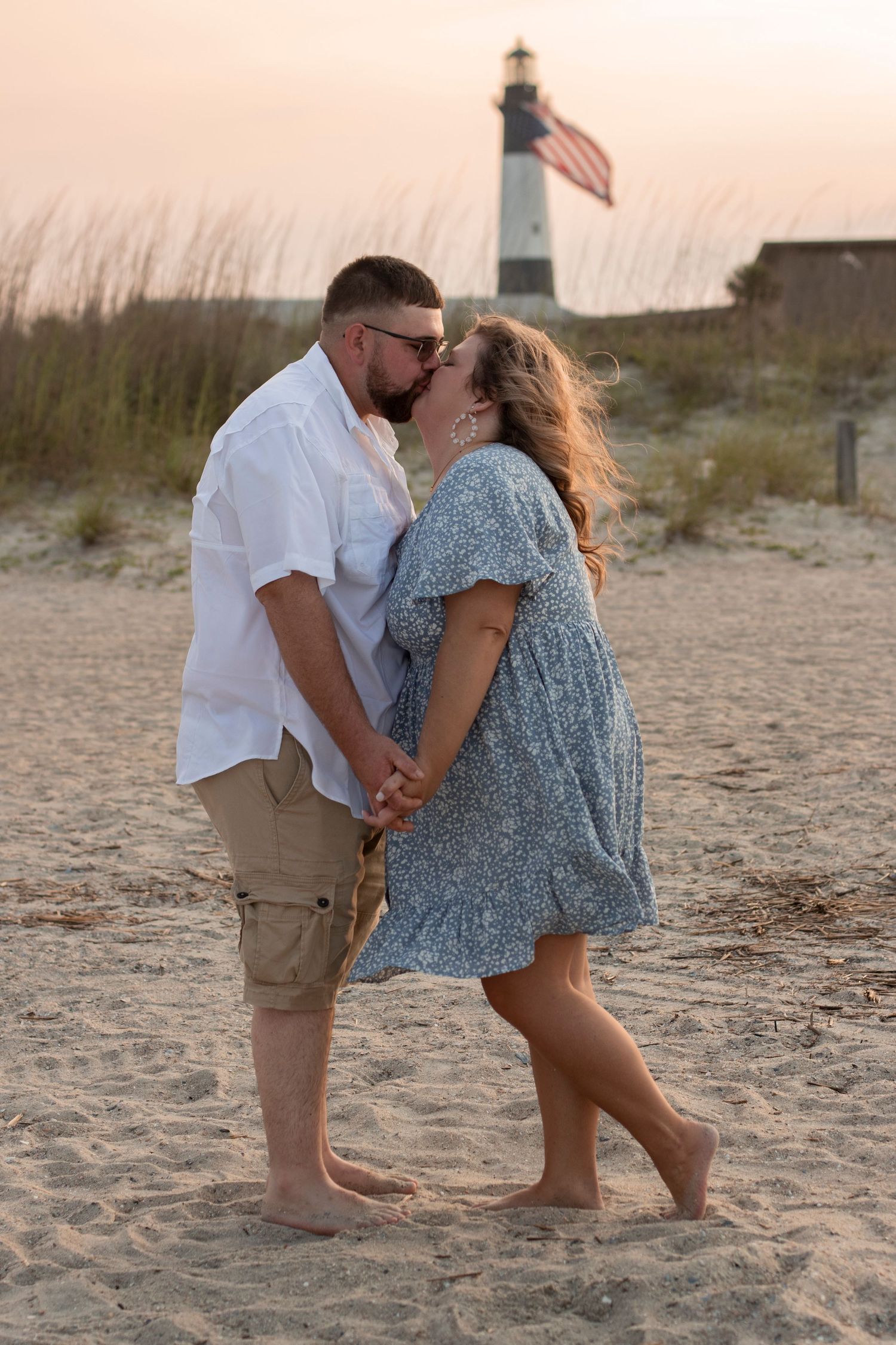 Romantic couple sharing tender moments on a beach with lighthouse in background at sunset.