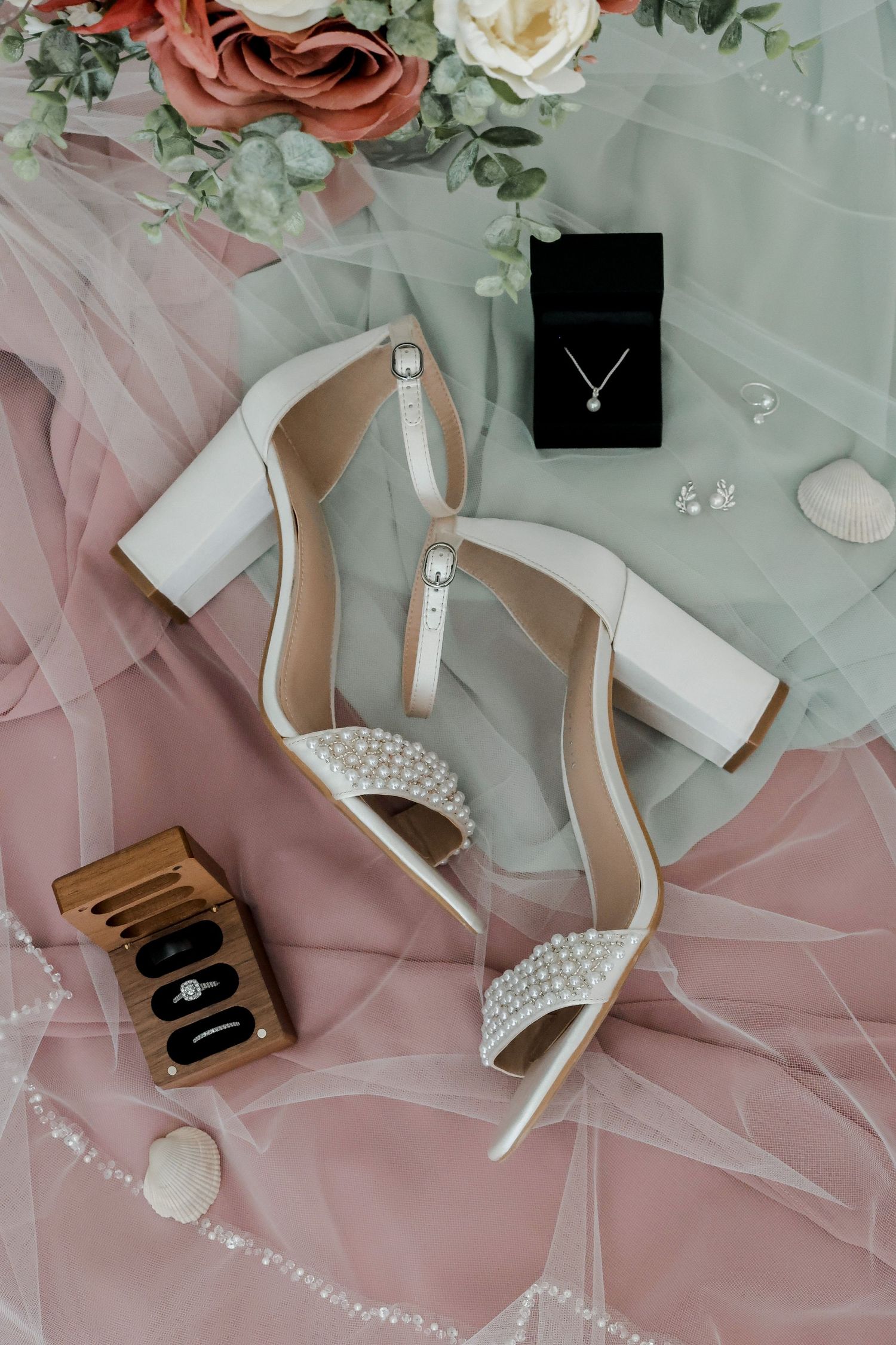 Wedding accessories including white heels, pearl necklace, and floral decorations on pink fabric.