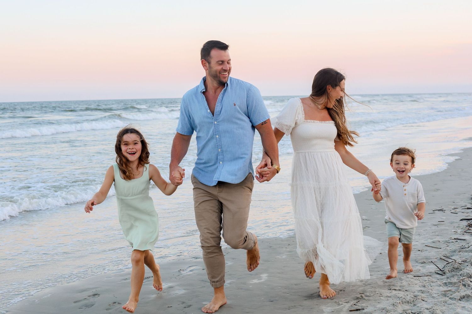 Family holding hands while running and playing together on a beach at sunset with ocean waves in background.