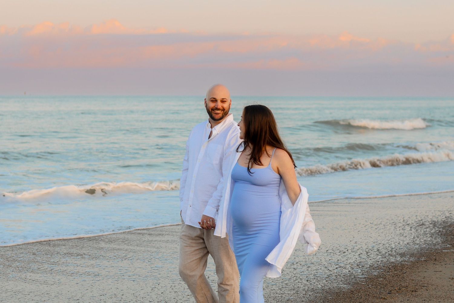 Couple walking along beach shoreline during pastel sunset.