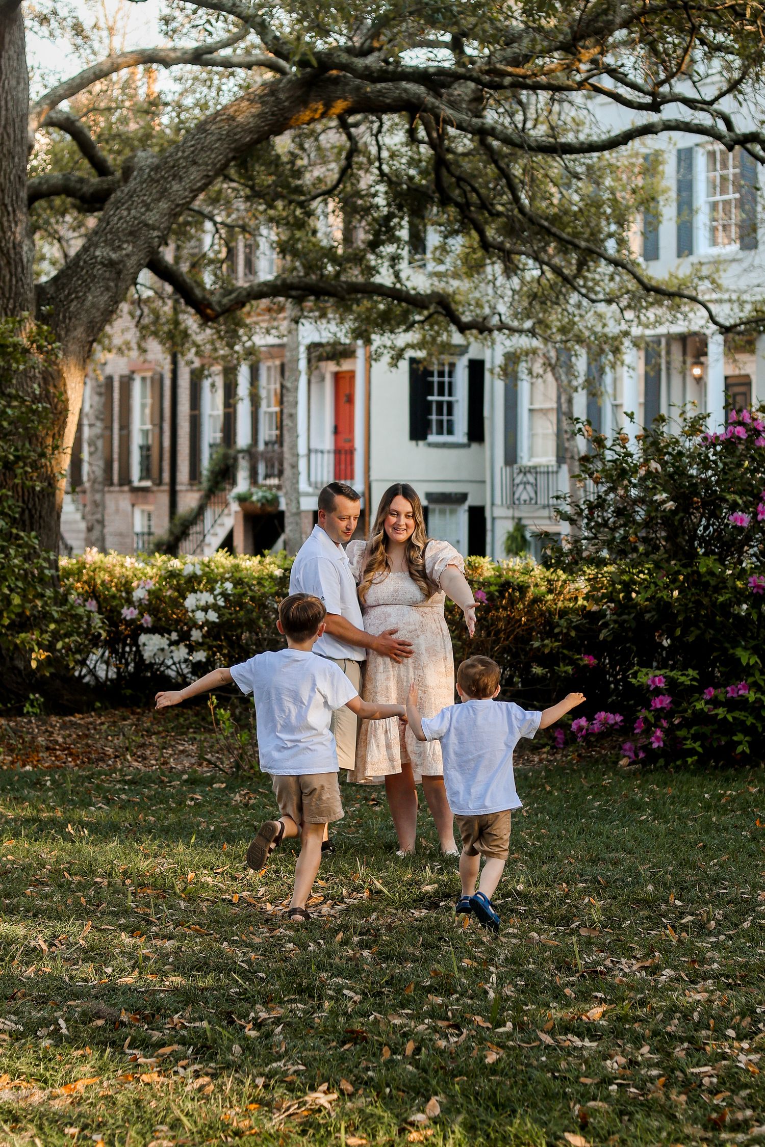 A family enjoying a sunny afternoon on a lawn in front of historic townhouses with moss-draped trees.