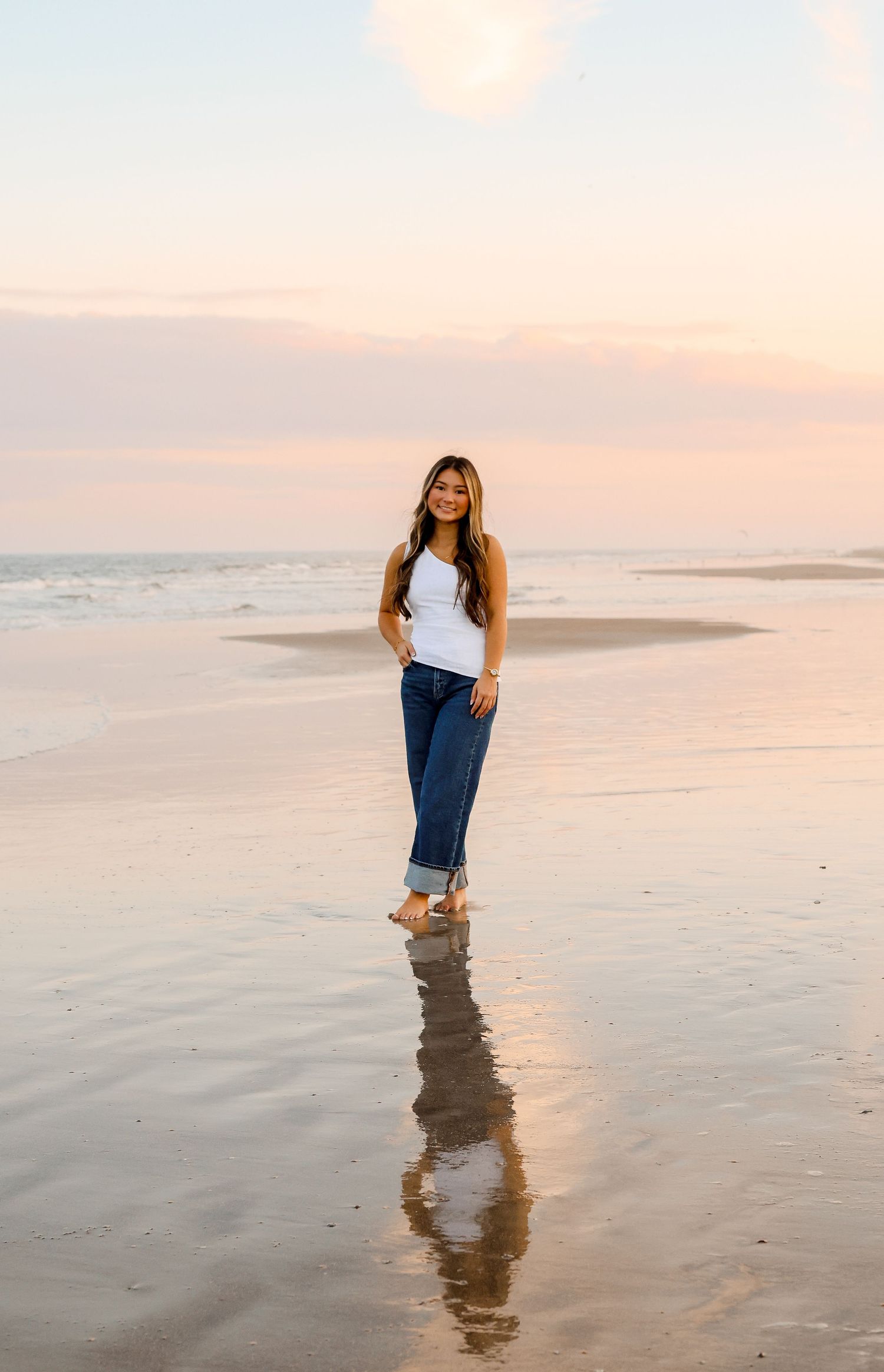 A person in jeans stands ankle-deep in shallow ocean water during sunset, creating a reflection on the wet sand.