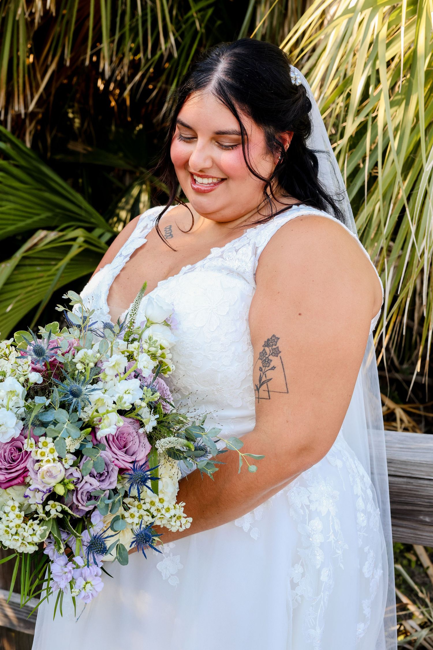 Wedding bouquet featuring purple roses and white hydrangeas held against flowing white bridal gown with palm trees in background.