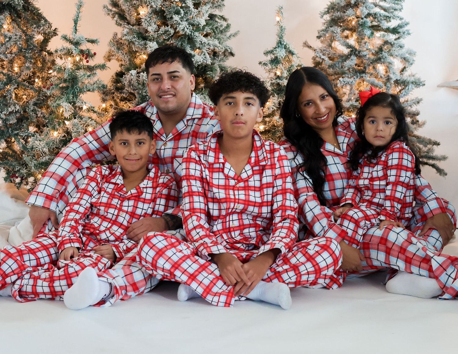 A family of five wearing matching red plaid pajamas poses in front of decorated Christmas trees.