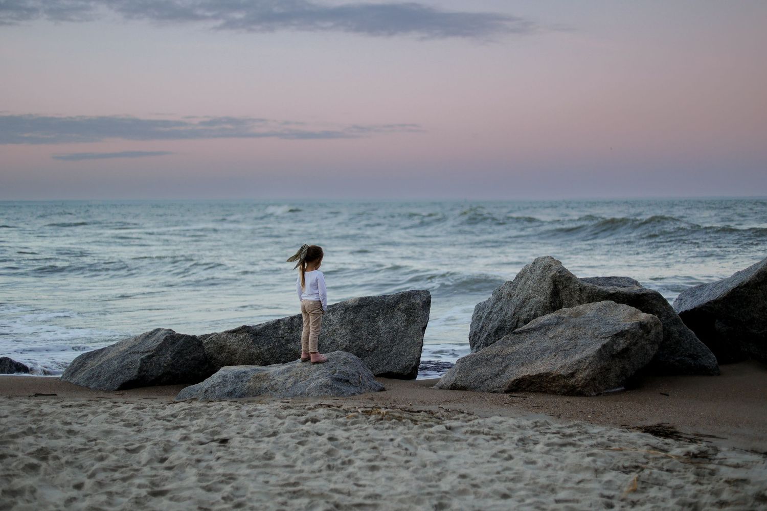 A silhouette stands on coastal rocks at sunset overlooking the ocean waves.