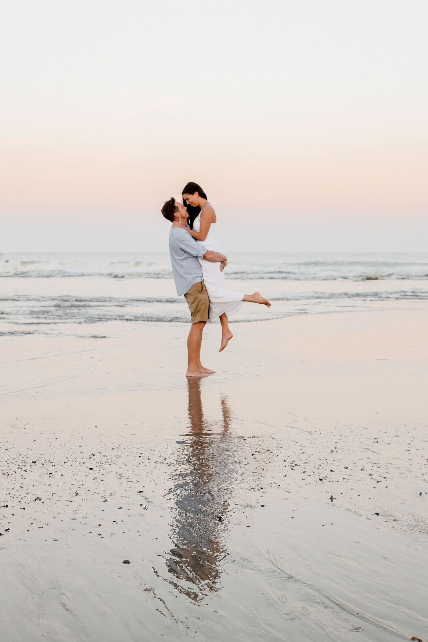 Couple embracing and lifting on a reflective beach at sunset creating a mirror image on wet sand.