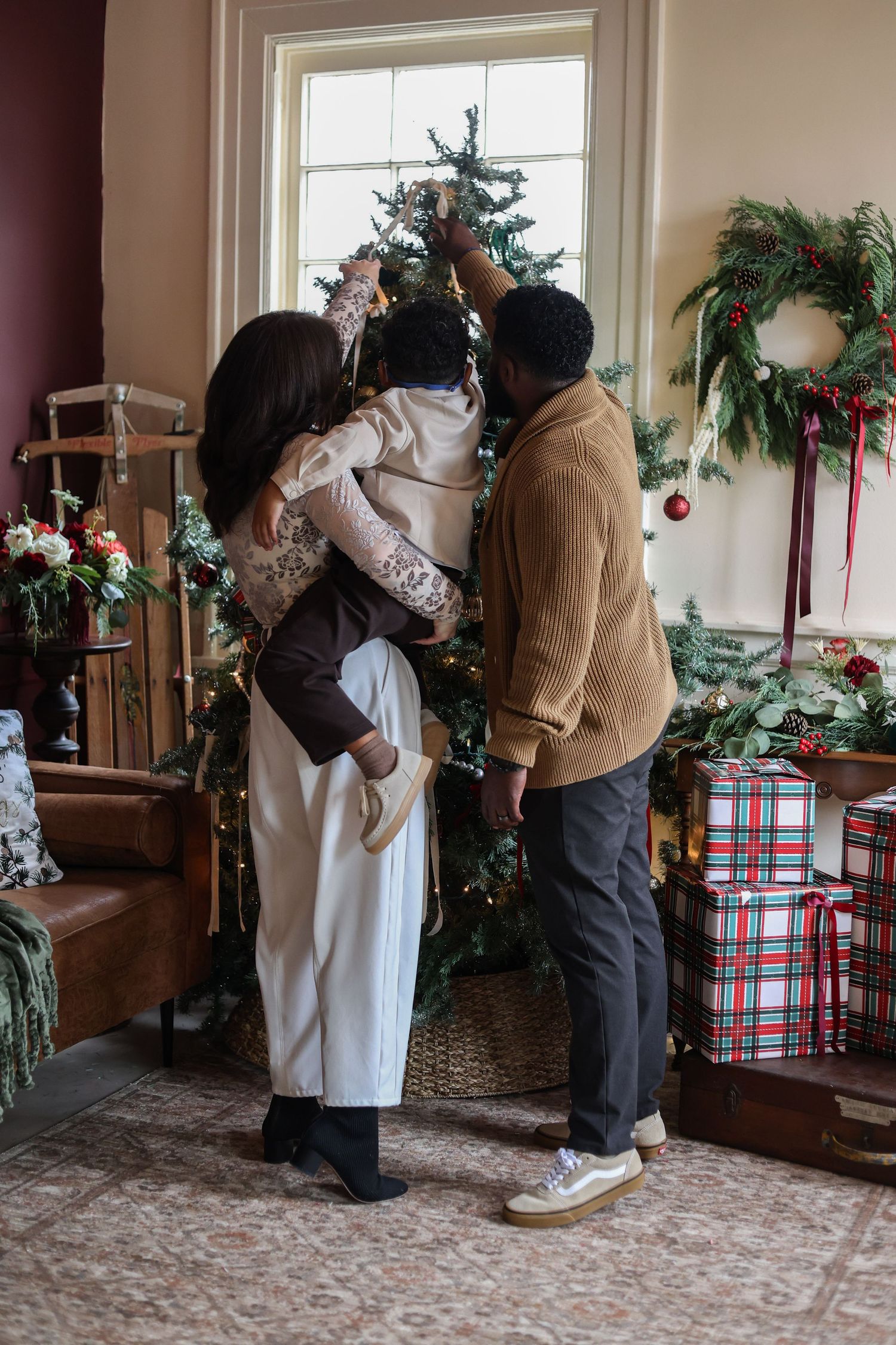 Family decorating a Christmas tree together as a woman lifts a child to place an ornament, with plaid gifts and a holly wreath nearby.
