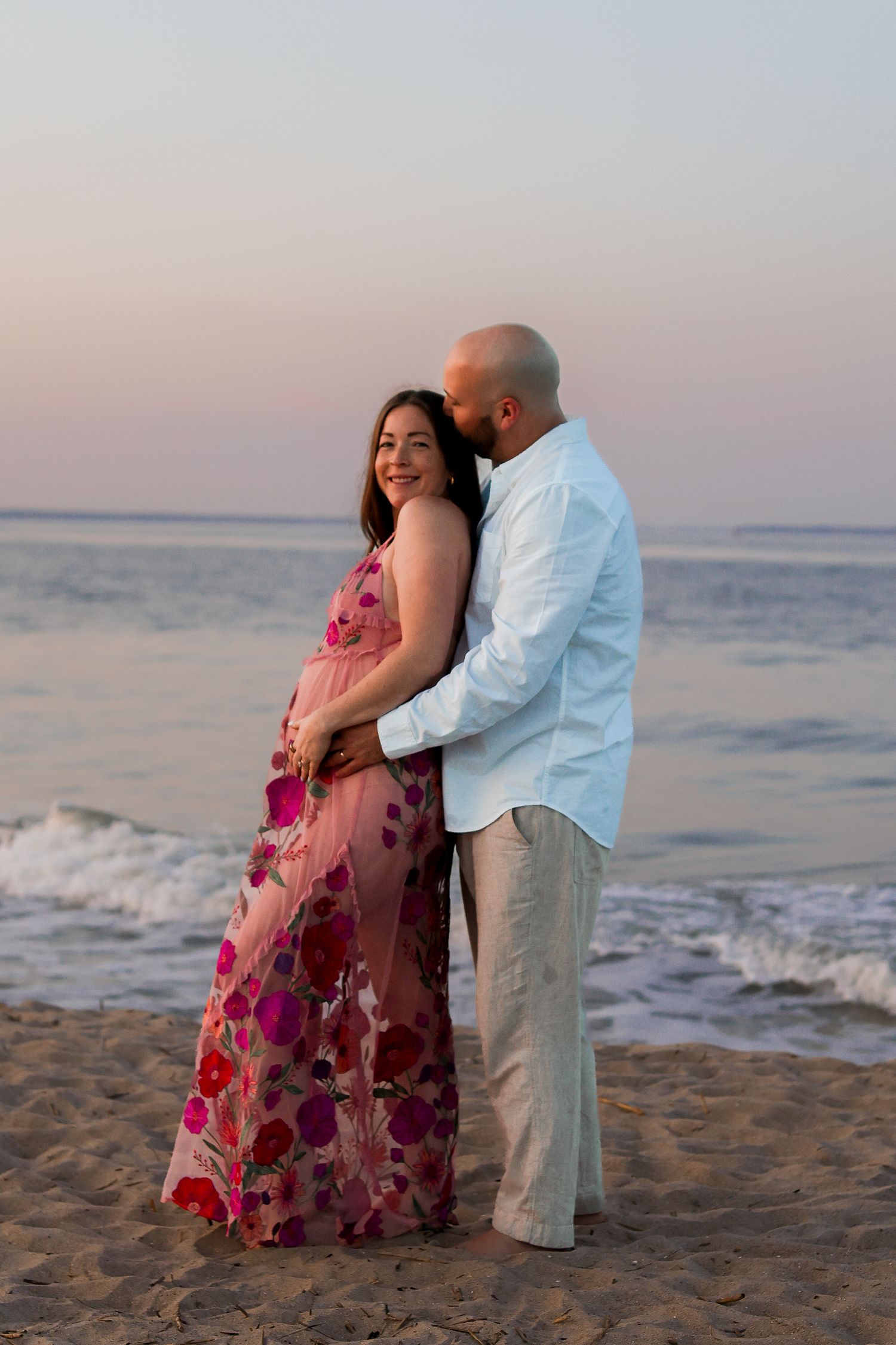 A romantic beach photoshoot at sunset with a couple in a pink floral dress and white shirt on sandy shores.
