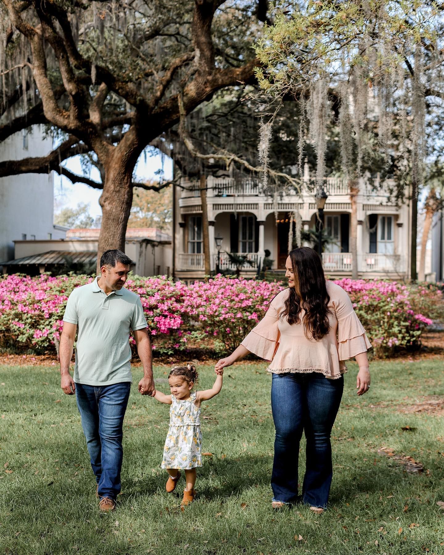 A family walks together on a grassy lawn with blooming azaleas and Spanish moss draped trees in the background.