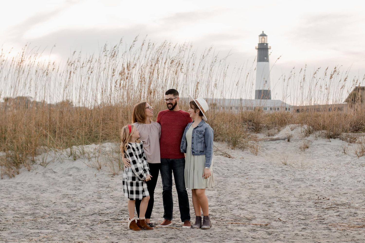 Family walks together on a sandy beach path through sea grass at sunset.