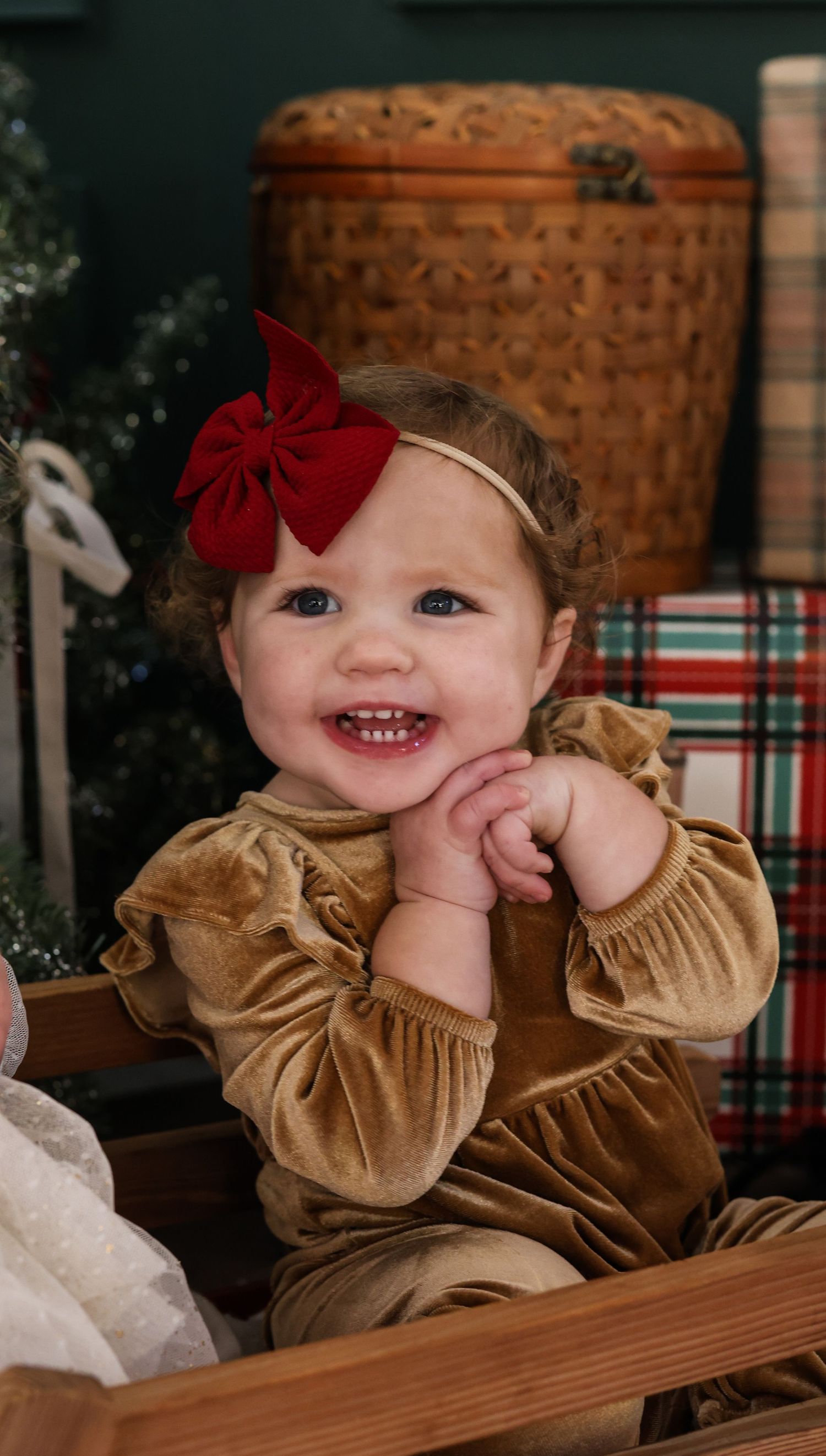 Smiling baby wearing gold velvet dress and red bow headband sits in wooden box with holiday decor in background.