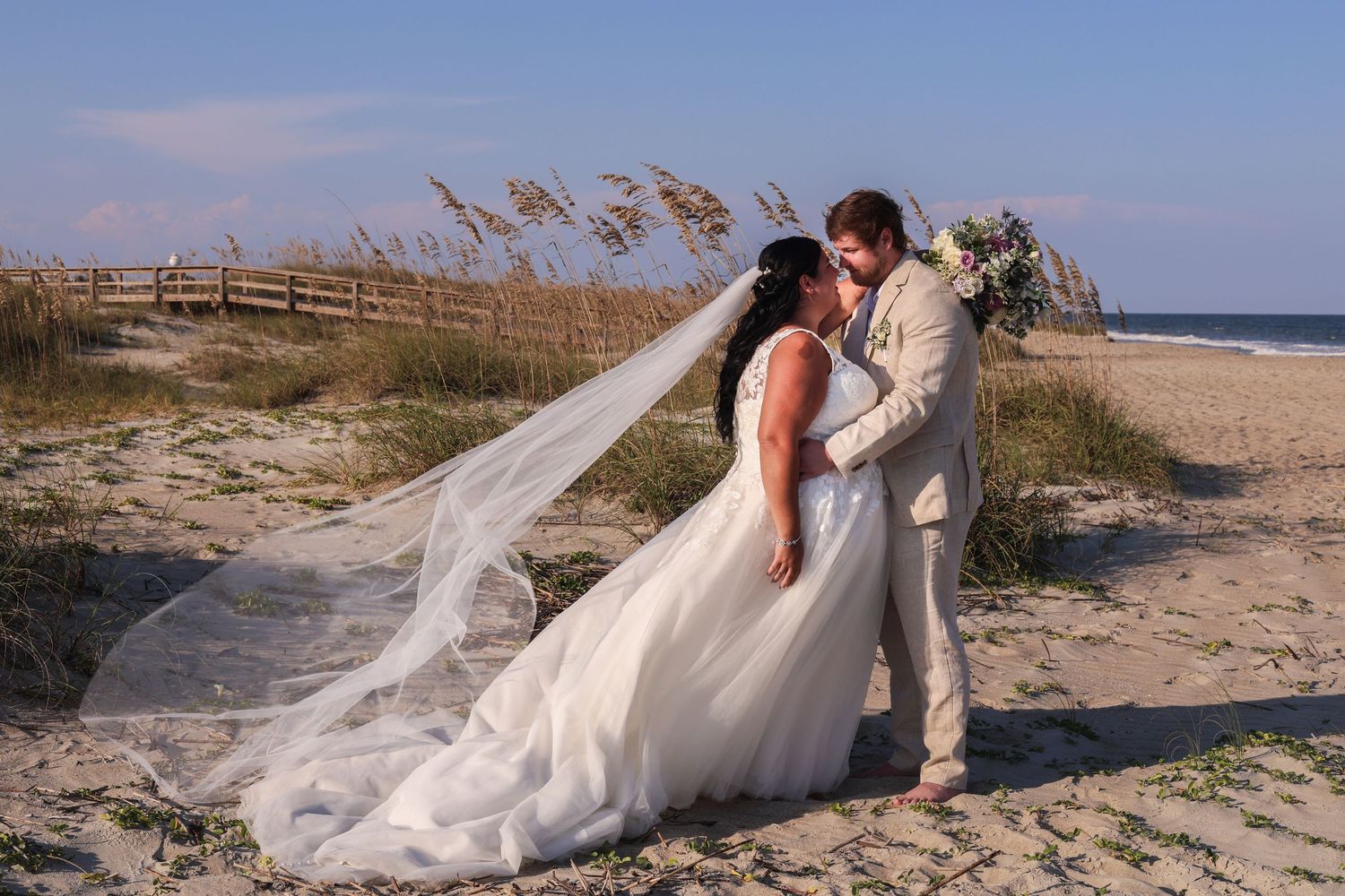 A couple in wedding attire walks along a sandy beach boardwalk with flowing veil in the wind at sunset.