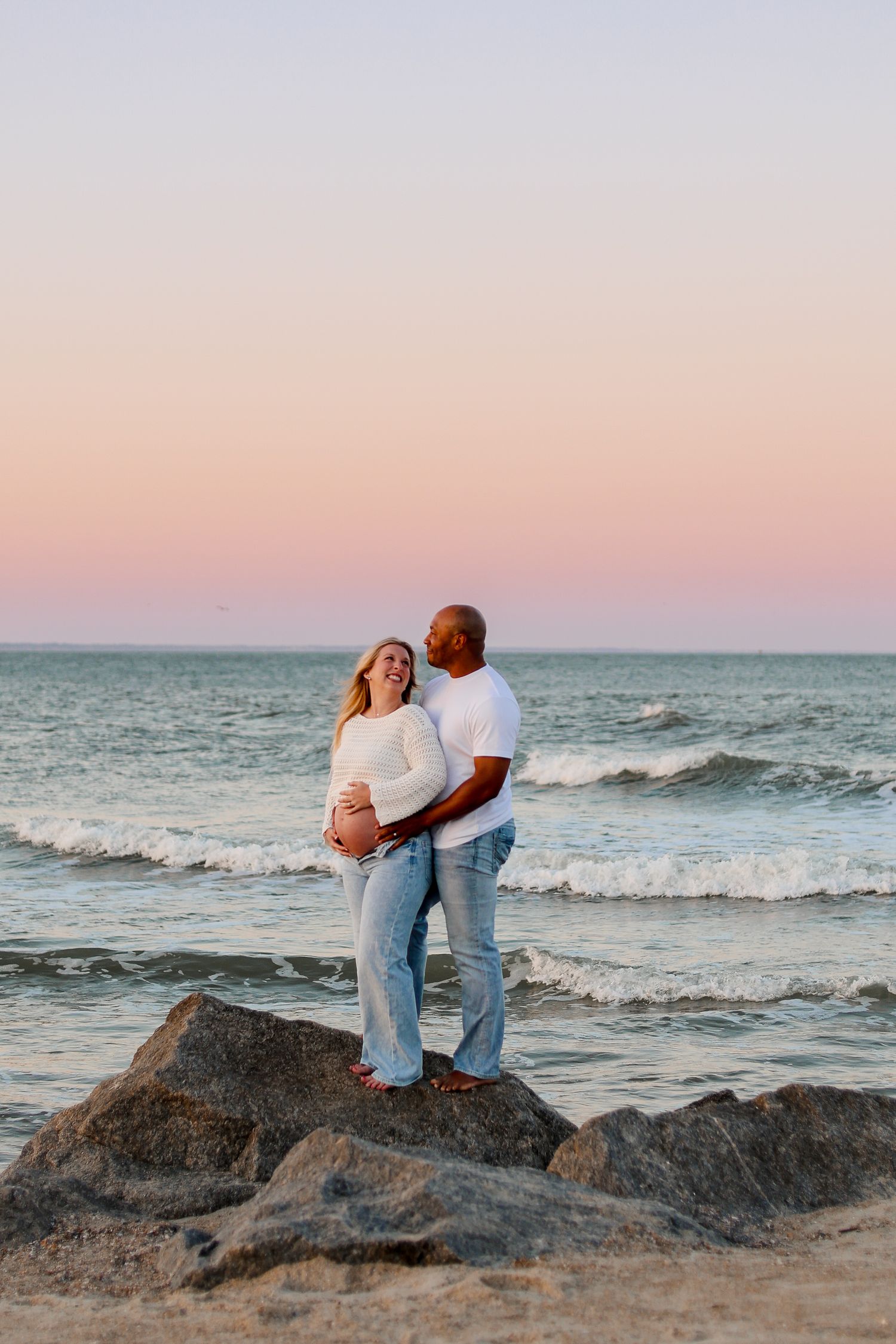 A couple shares tender moments on beach rocks during sunset with waves in the background.