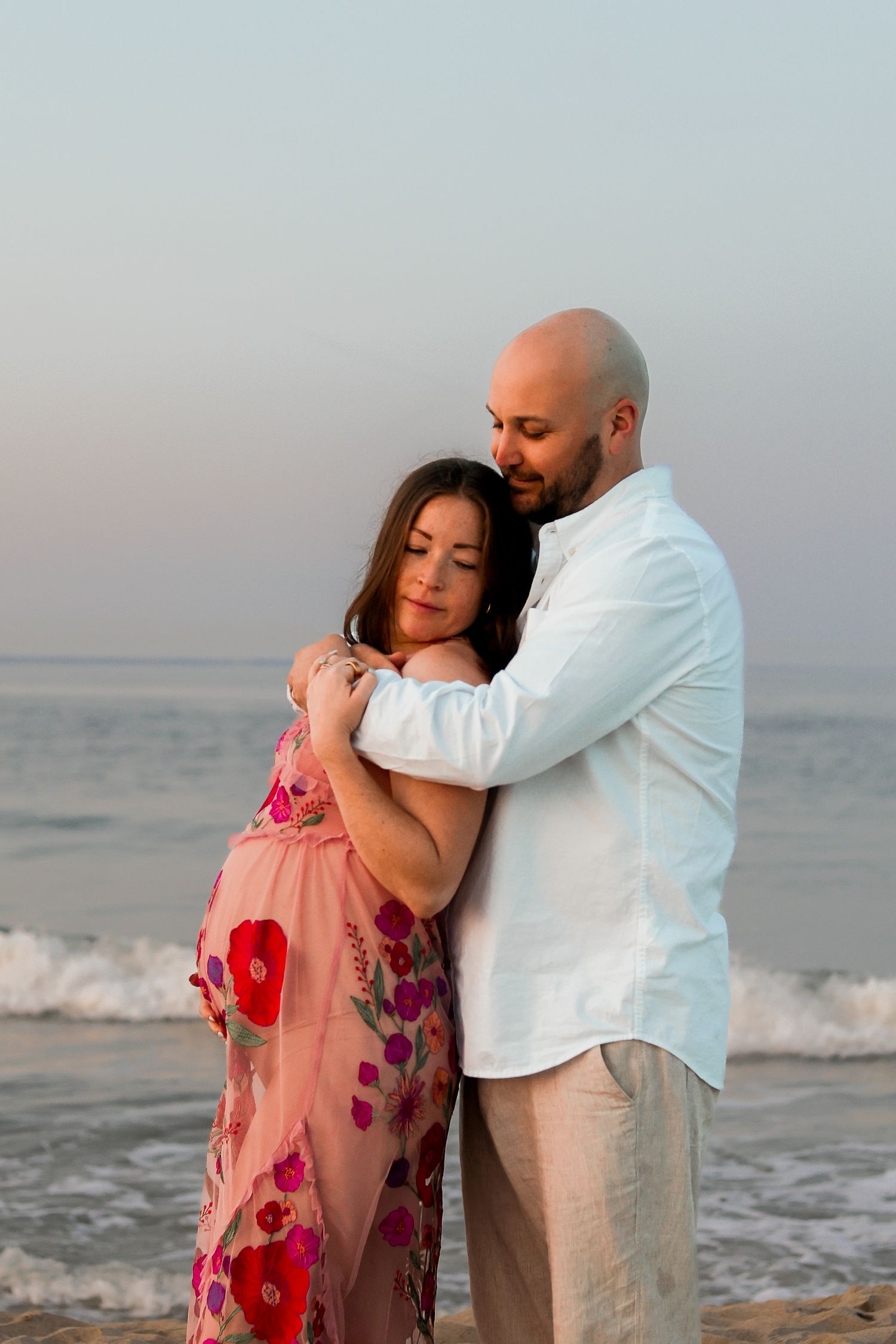 A couple embraces tenderly on a beach at sunset, with the woman wearing a floral pink dress and man in white shirt.