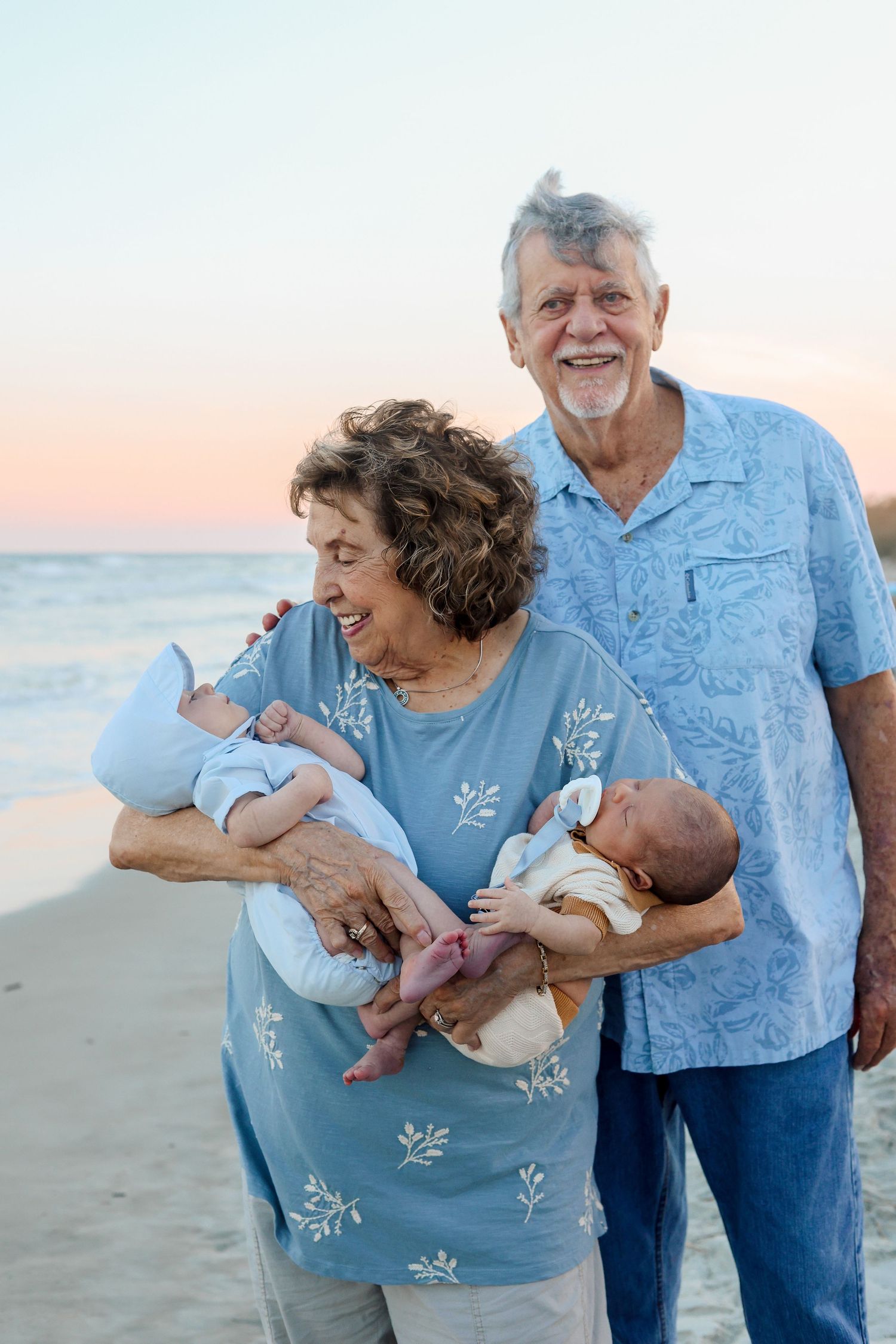 A couple in blue outfits holds twin babies while standing on a beach at sunset.