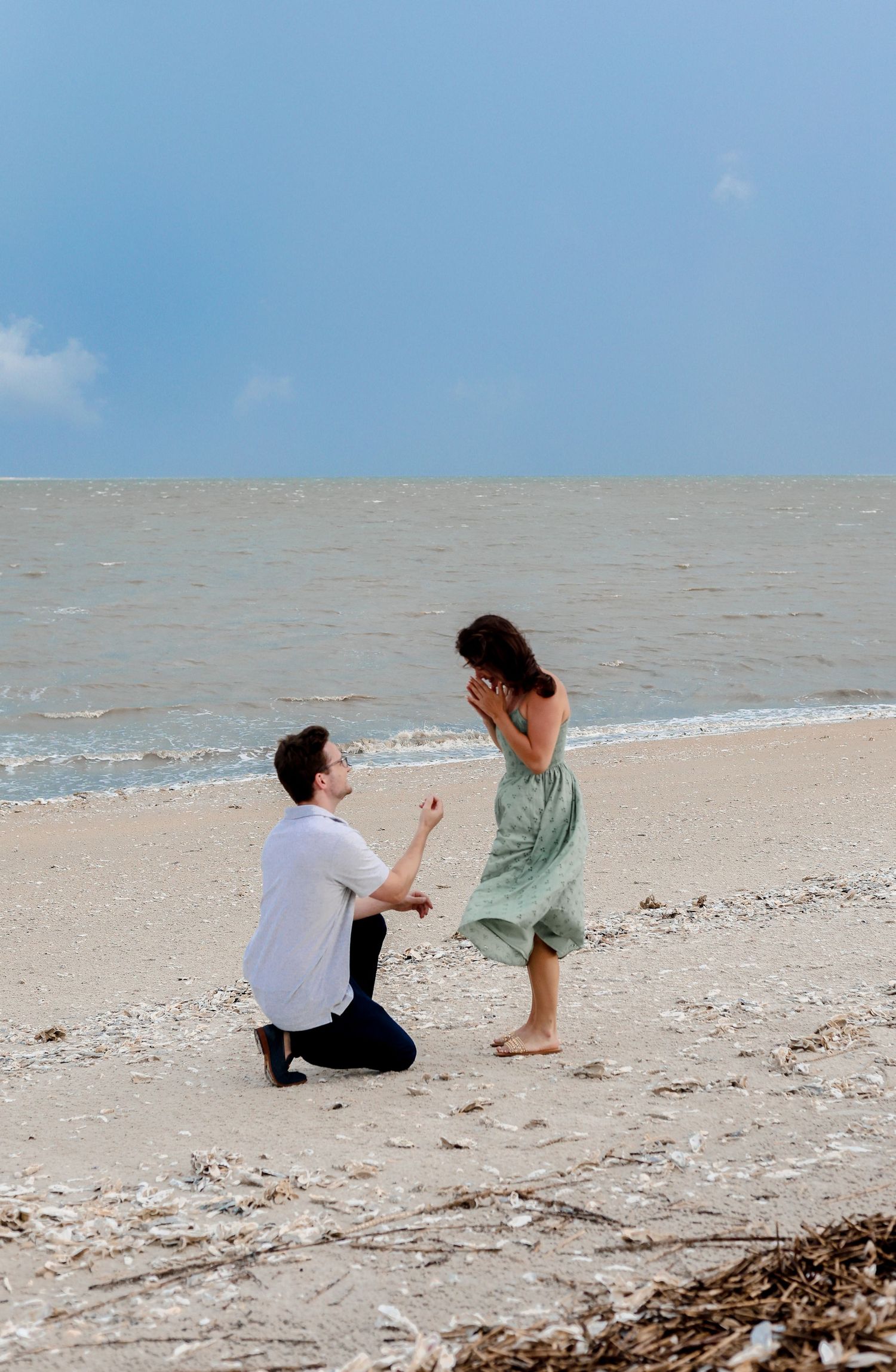 A romantic beach proposal scene with someone kneeling in the sand as waves crash on the shore under a blue sky.