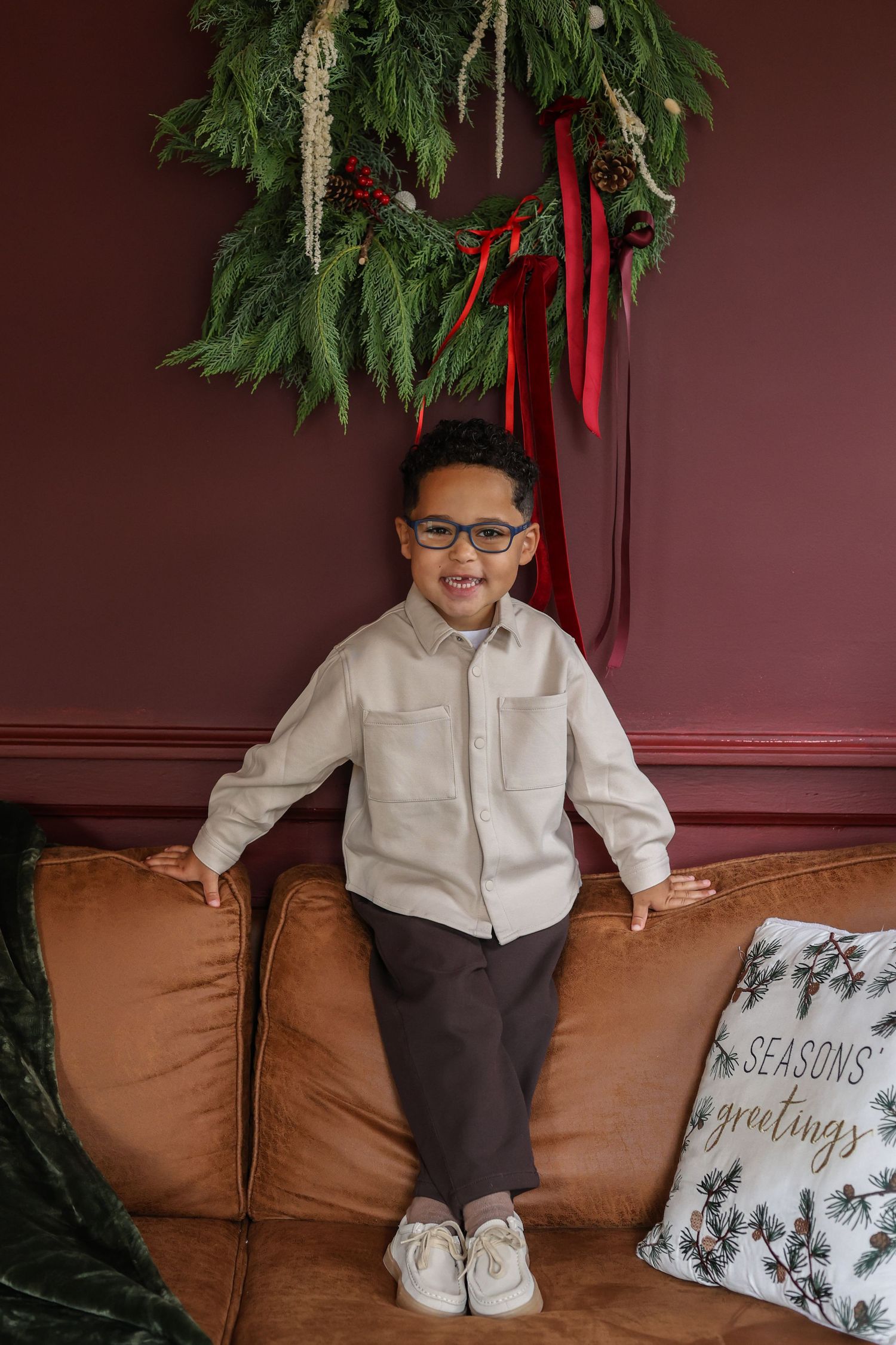 Smiling boy in glasses and a cream button-up shirt stands on a tan leather couch beneath a red-ribboned evergreen holiday wreath.