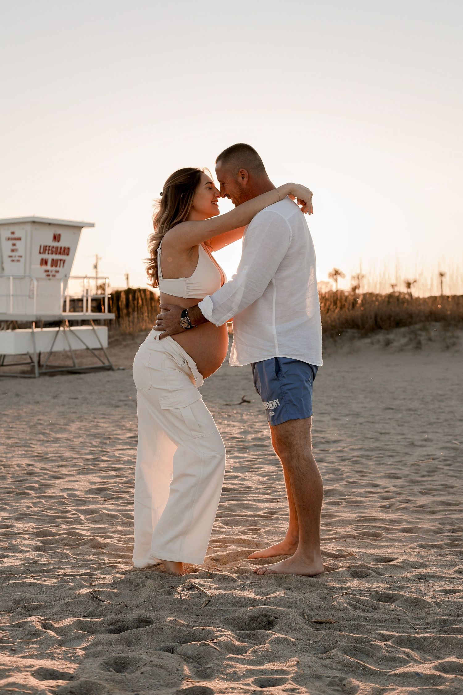 Romantic couple sharing intimate moments on the beach at sunset near a lifeguard tower.