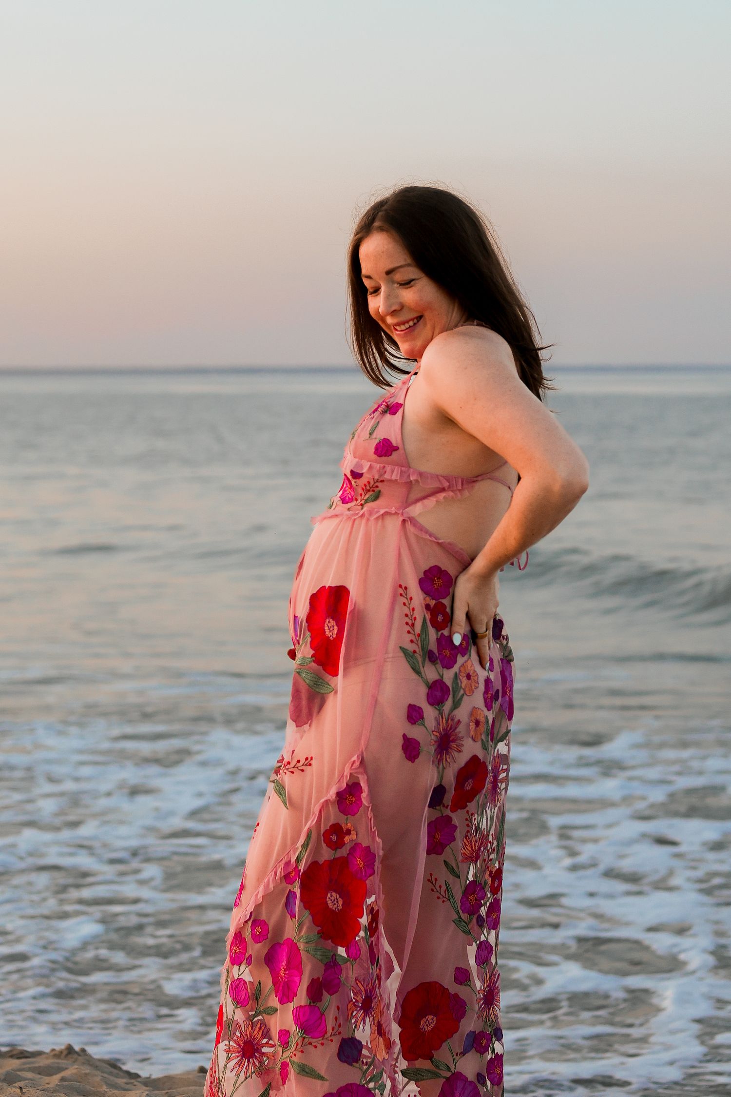 Happy mom-to-be in floral dress laughs on beach at sunset.