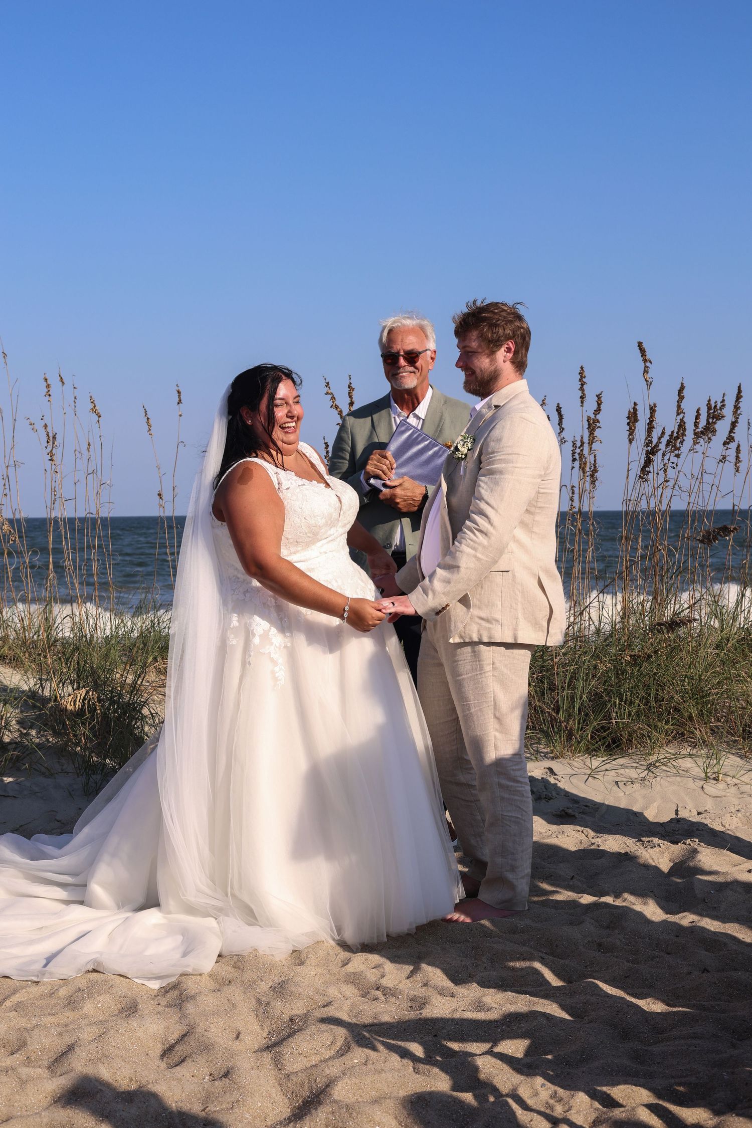 A wedding ceremony takes place on a sandy beach with sea oats and blue skies in the background.