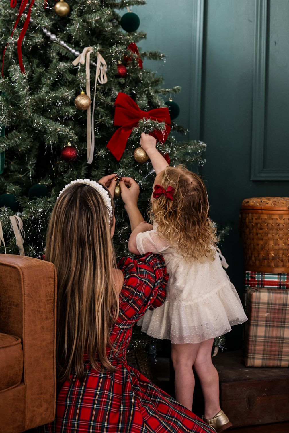 Two figures in red plaid and white dress decorating a Christmas tree together against a teal wall.
