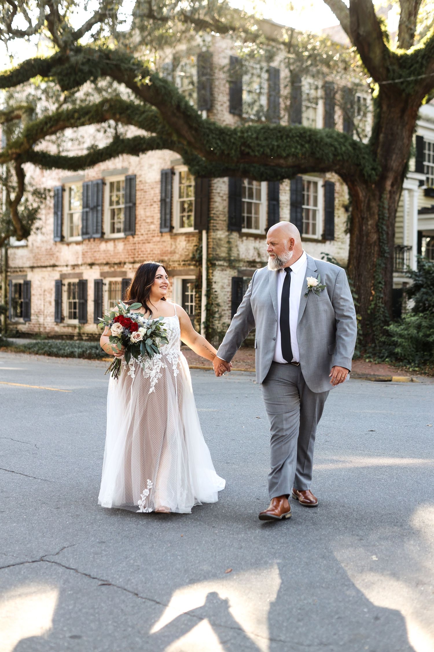 Wedding couple walks hand in hand under majestic oak trees in a historic courtyard.