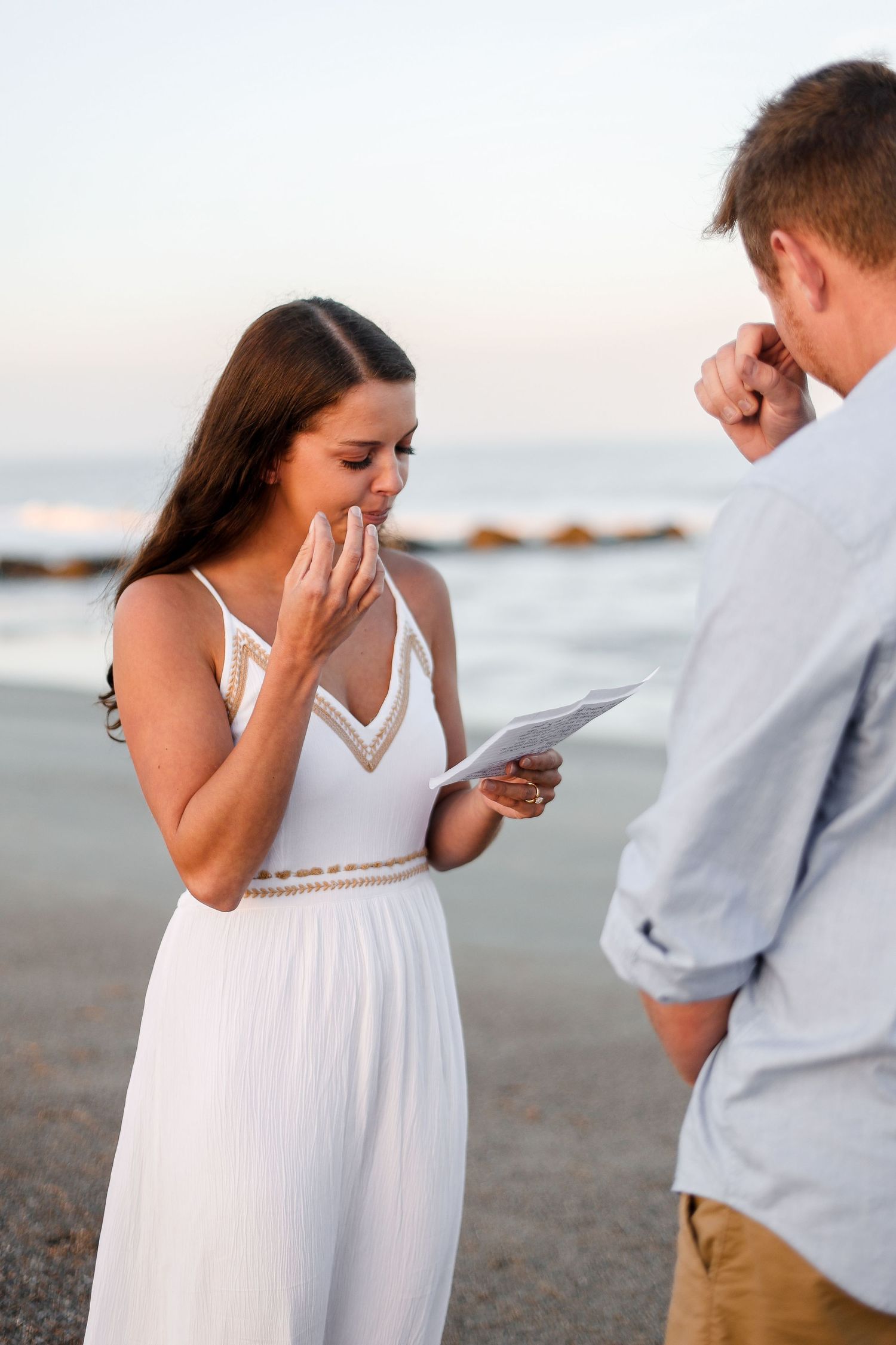 A couple shares an emotional moment during a surprise beach proposal as the person in a white dress reads a letter.