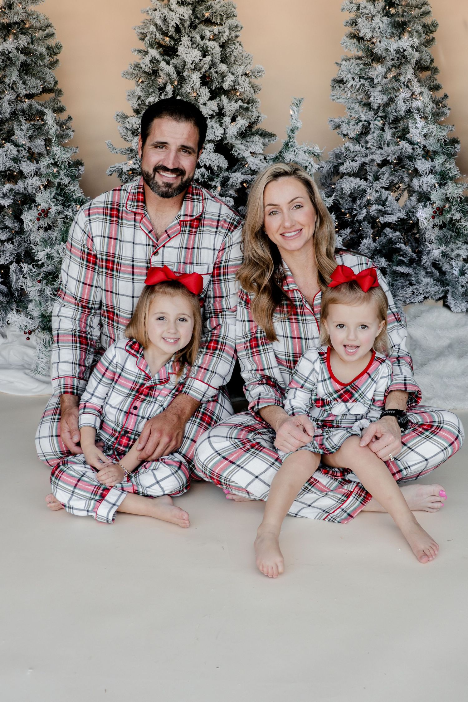 Family wearing matching plaid Christmas pajamas posing in front of snowy evergreen trees.