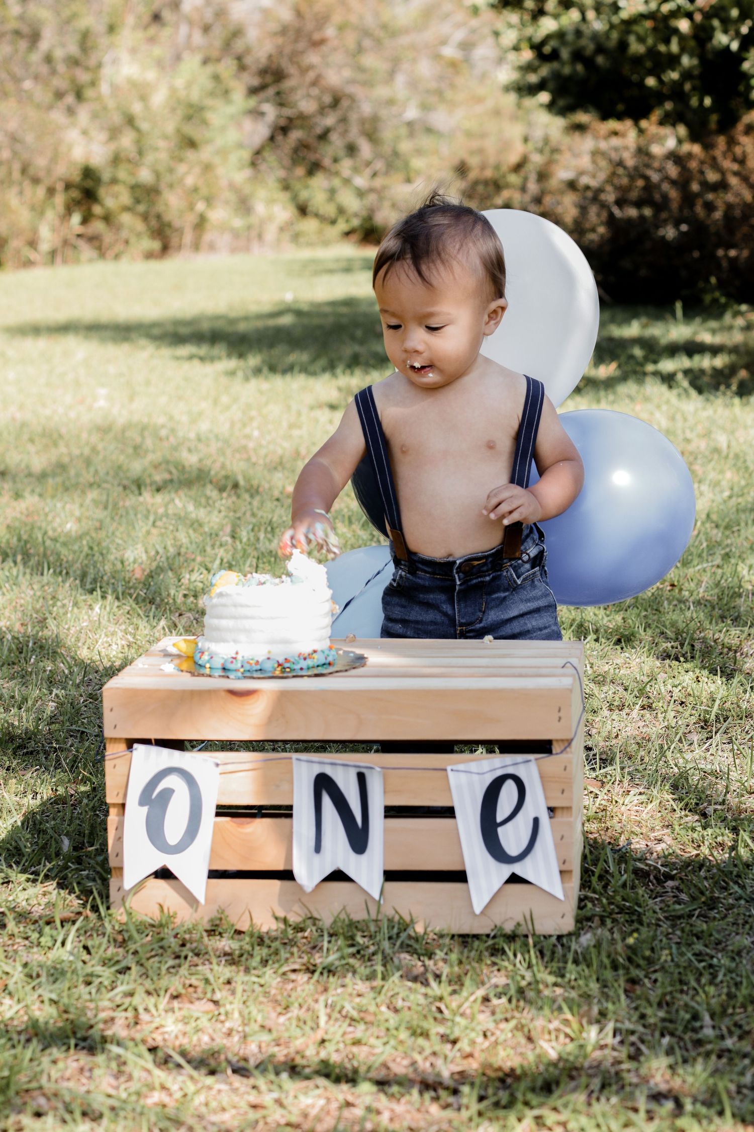 Baby enjoying cake smash session on wooden crate outdoors with birthday decorations.