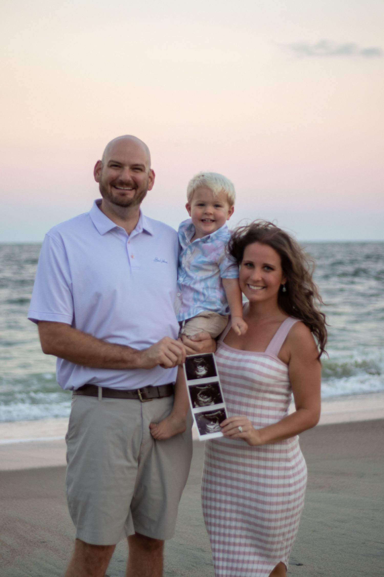 A couple and child hold ultrasound photos while posing on a sunset beach, showing their pregnancy announcement.