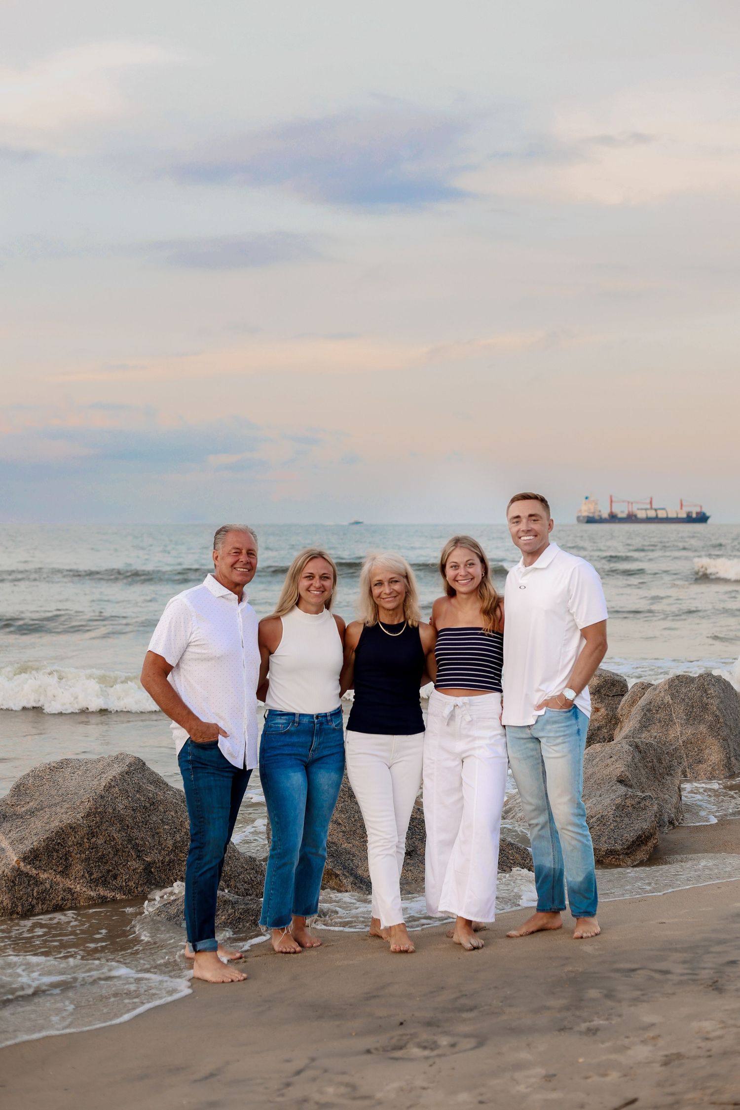 Family group poses together on a beach at sunset with ocean waves and rocks in the background.
