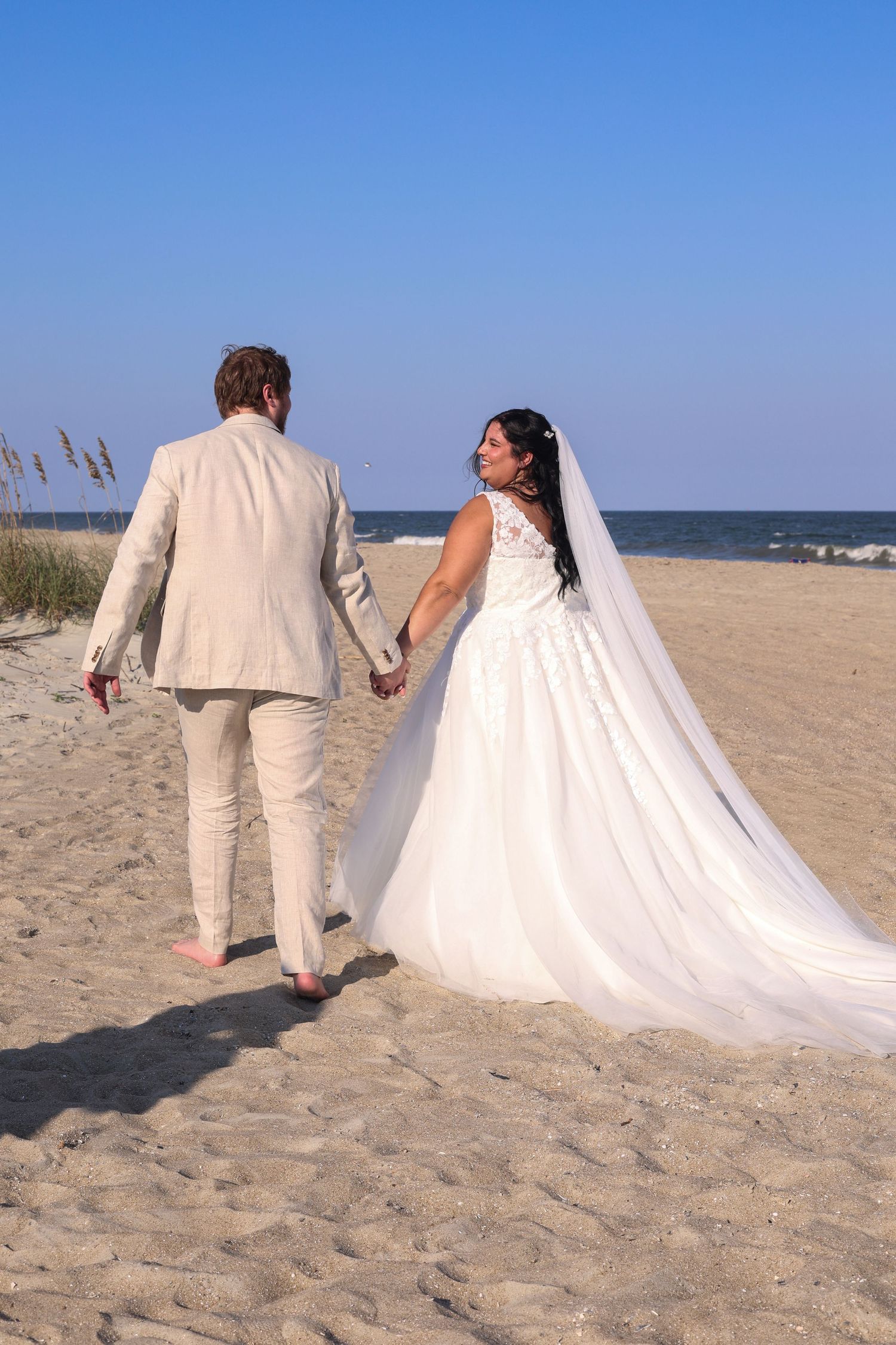 A couple in wedding attire walks along a sandy beach with ocean waves in the background.