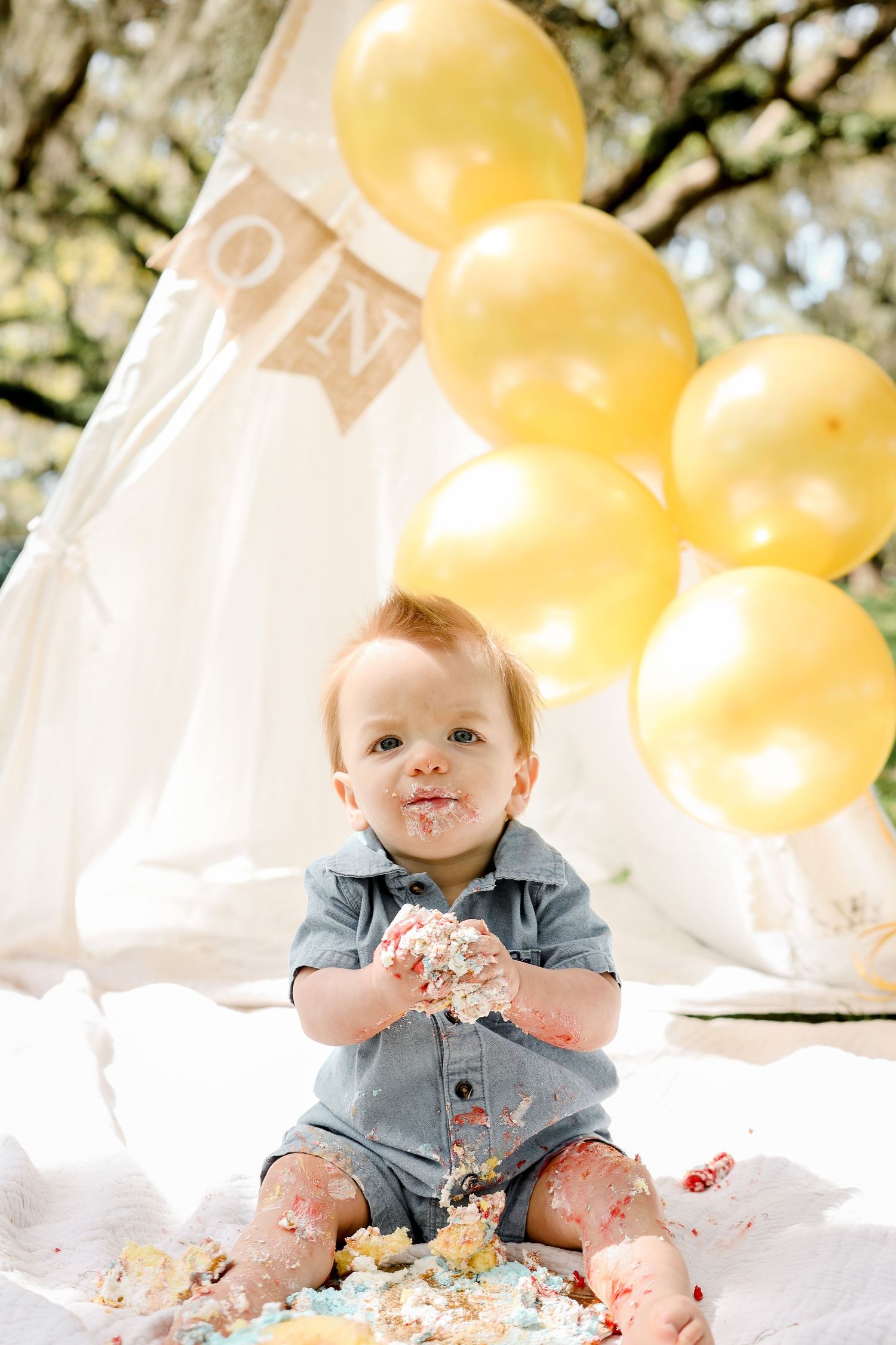 Cake smash photo with white teepee, yellow balloons and ONE banner in background.