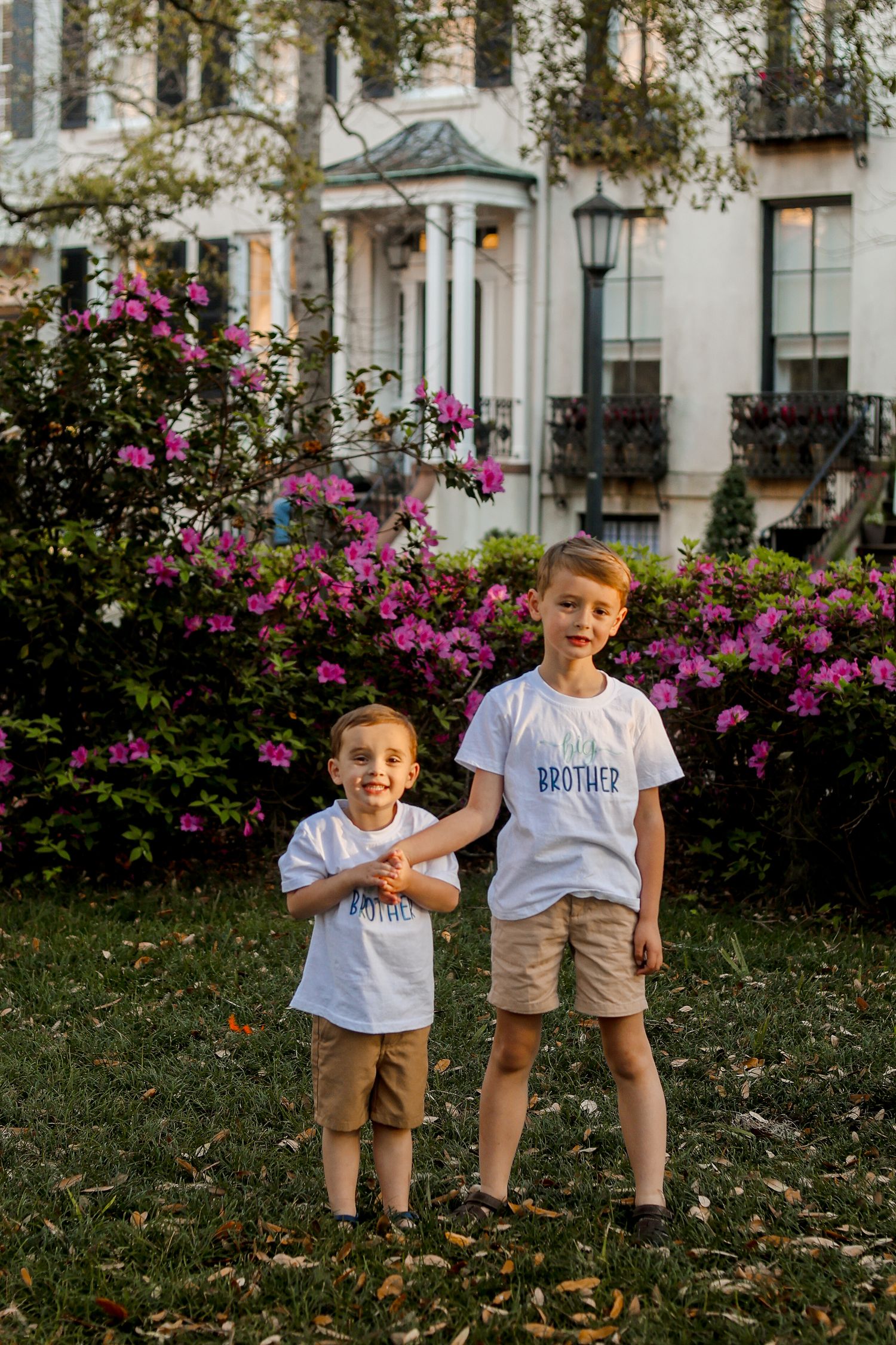 Two young children posing in matching outfits by blooming azalea bushes.