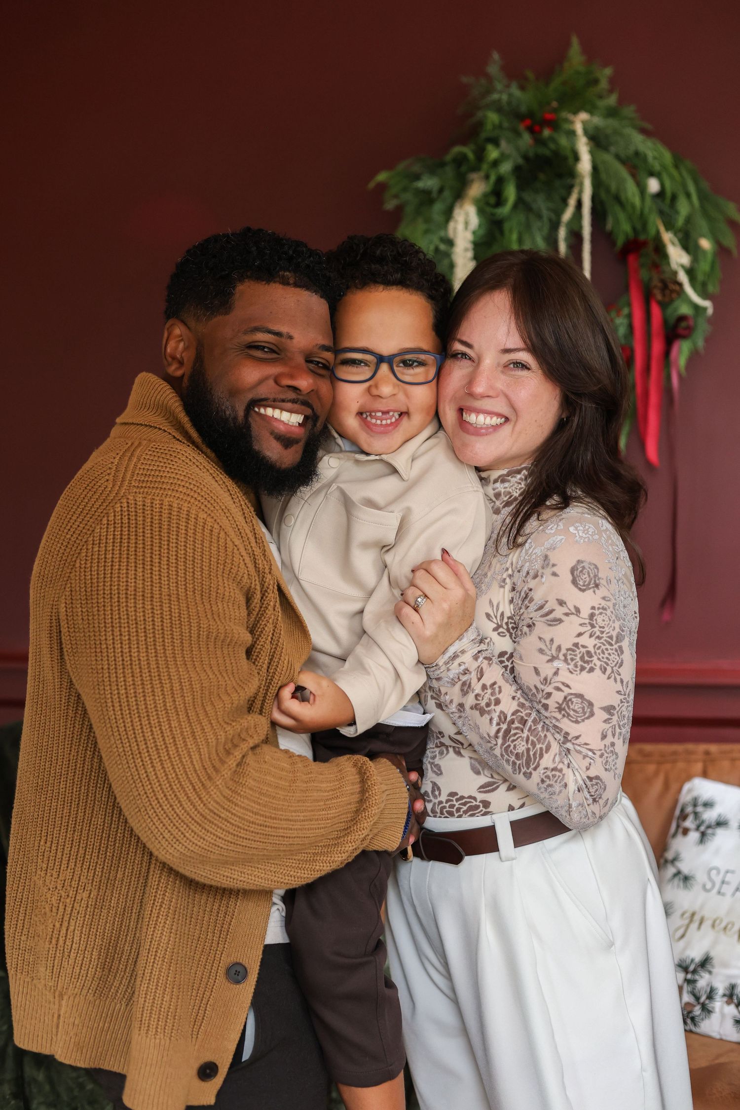 A smiling man, woman, and young boy in glasses hug closely in a festive family holiday portrait with a evergreen wreath on a burgundy wall.