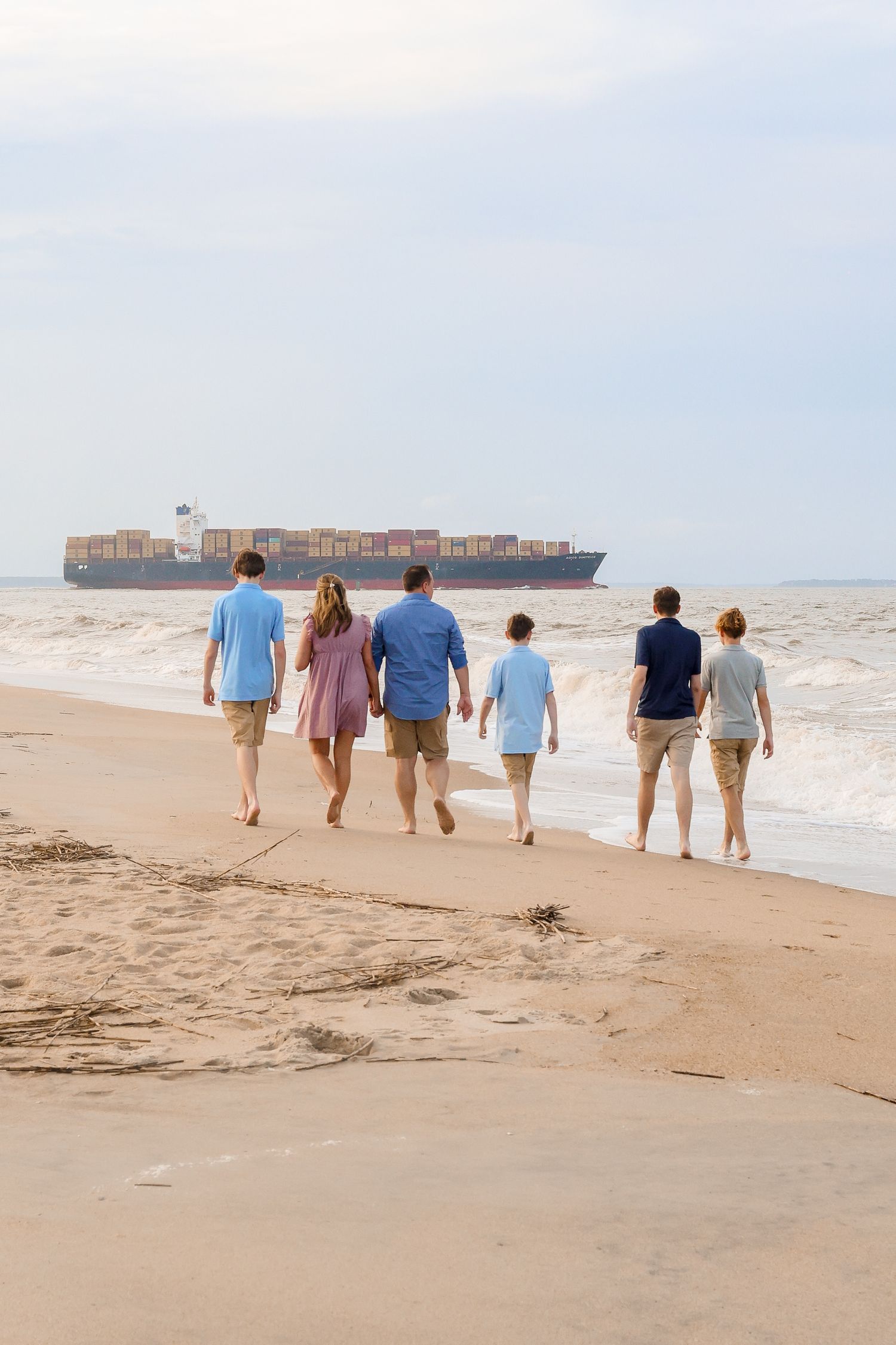 Group of people walking along sandy beach at sunset with large cargo ship visible in background across the horizon.