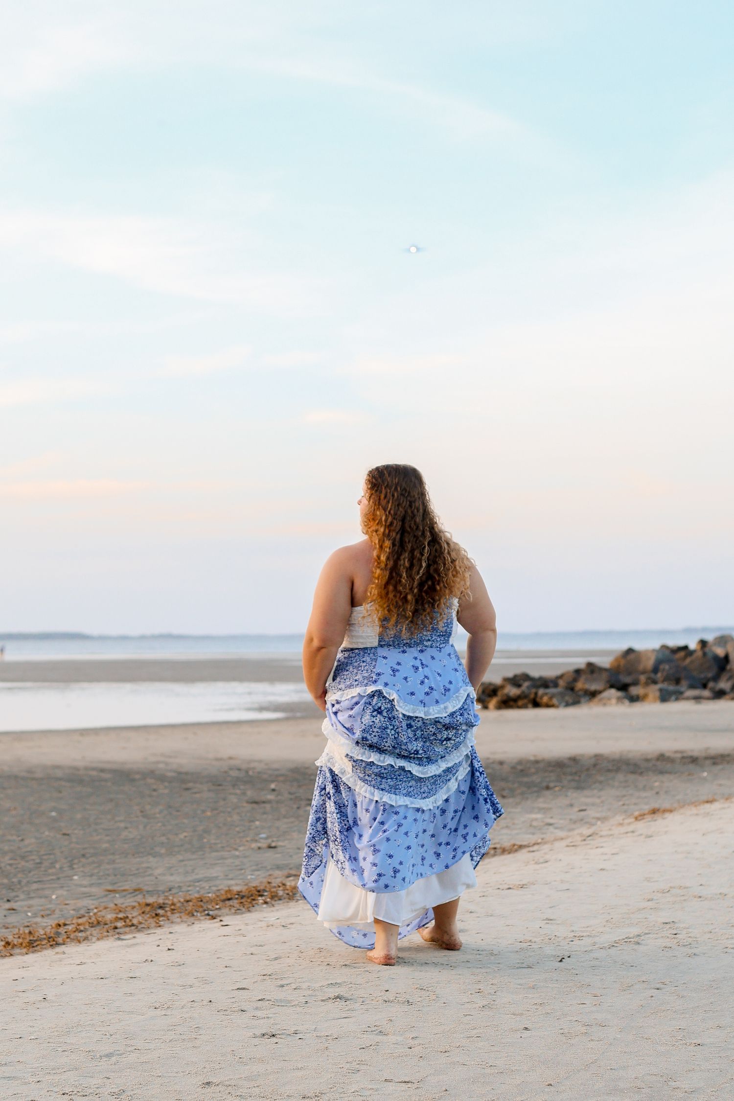 Person in flowing blue and white patterned dress walks barefoot along a peaceful beach at sunset.